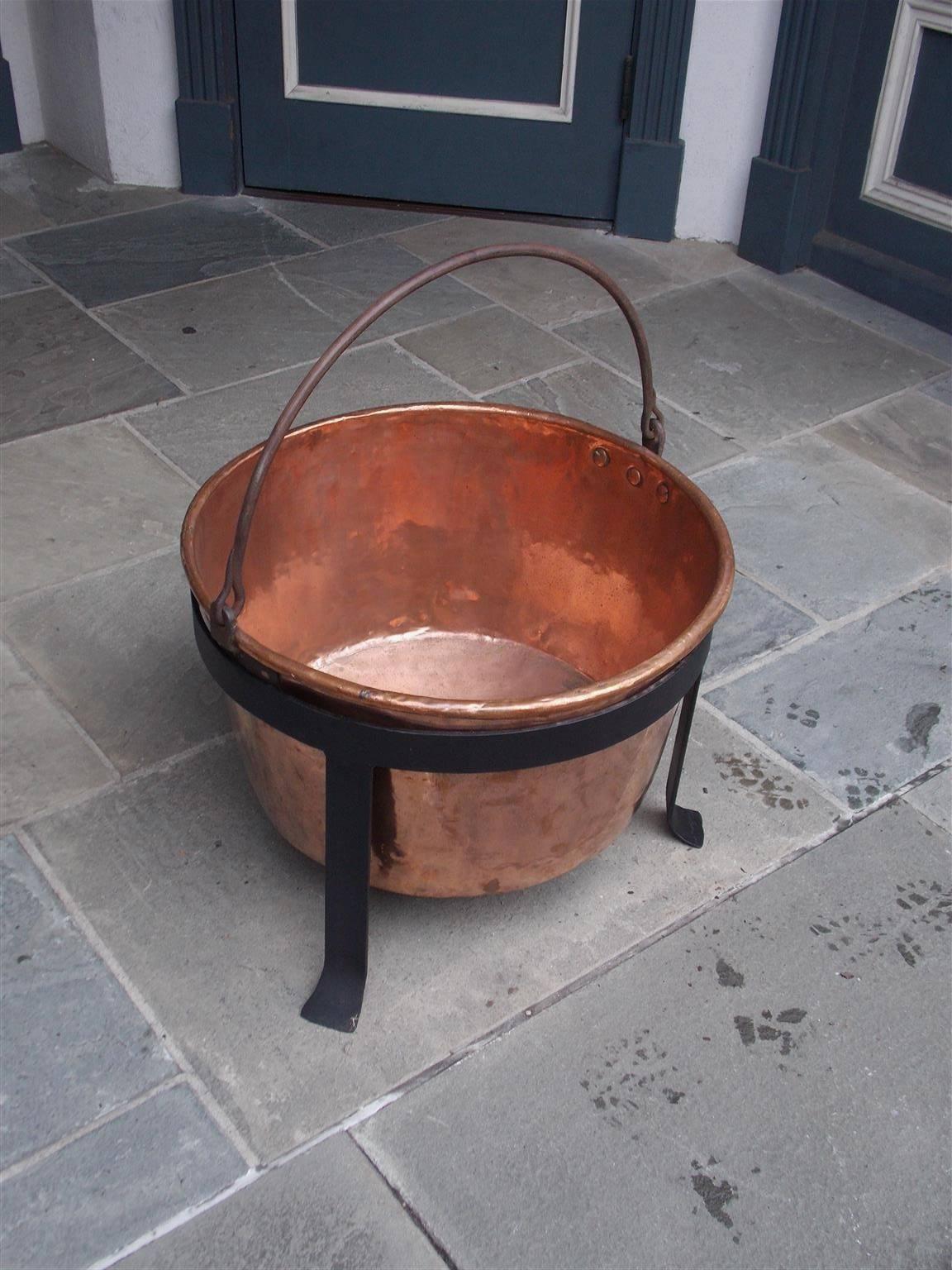 American copper and wrought iron plantation cauldron with original iron folding handle supported by circular fitted three leg stand with stylized splayed feet. Late 18th Century.