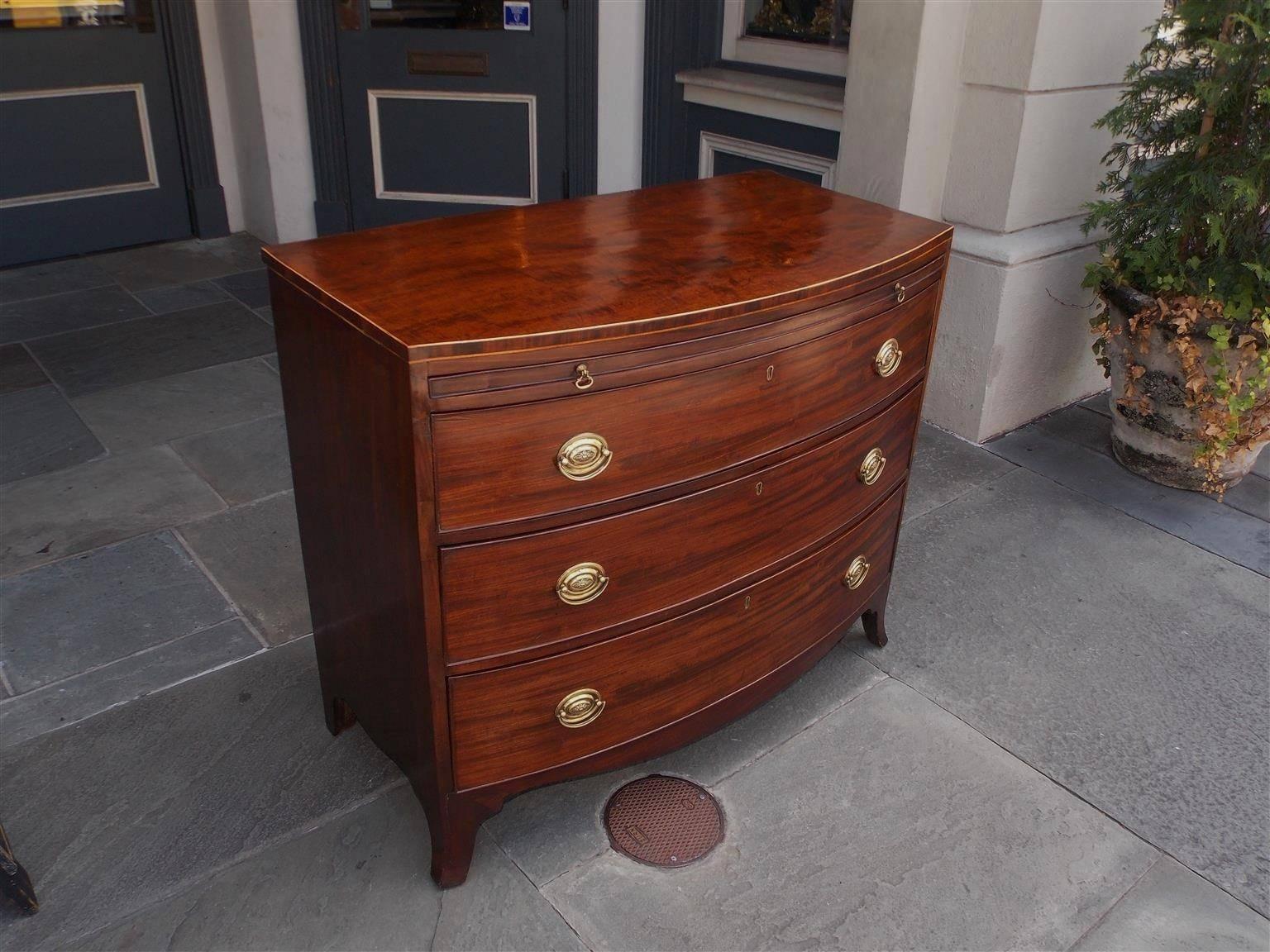 English mahogany bow front graduated three-drawer chest with satinwood string inlay, upper baize brushing slide, period brasses, carved scalloped skirt, and terminating on the original splayed French feet,  Late 18th century.
