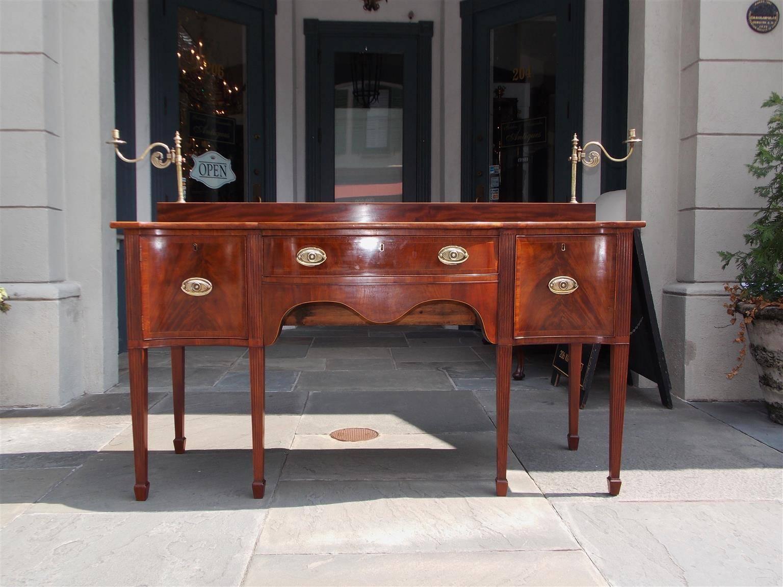 American mahogany reverse serpentine satinwood inlaid sideboard with a stage top having flanking adjustable brass single candelabras, a center drawer flanked by cabinet and wine drawer, original brasses, a carved serpentine molded skirt and