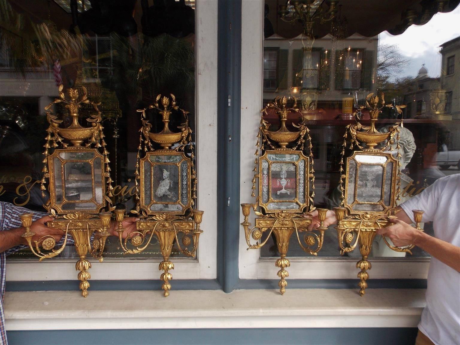 Set of Four English George III Adam gilt carved wood two arm wall sconces with centered urns, flanking graduated bell flowers, acid etched mirrors, scrolled floral medallions, and terminating with tiered graduated bell flowers. Late 18th Century.