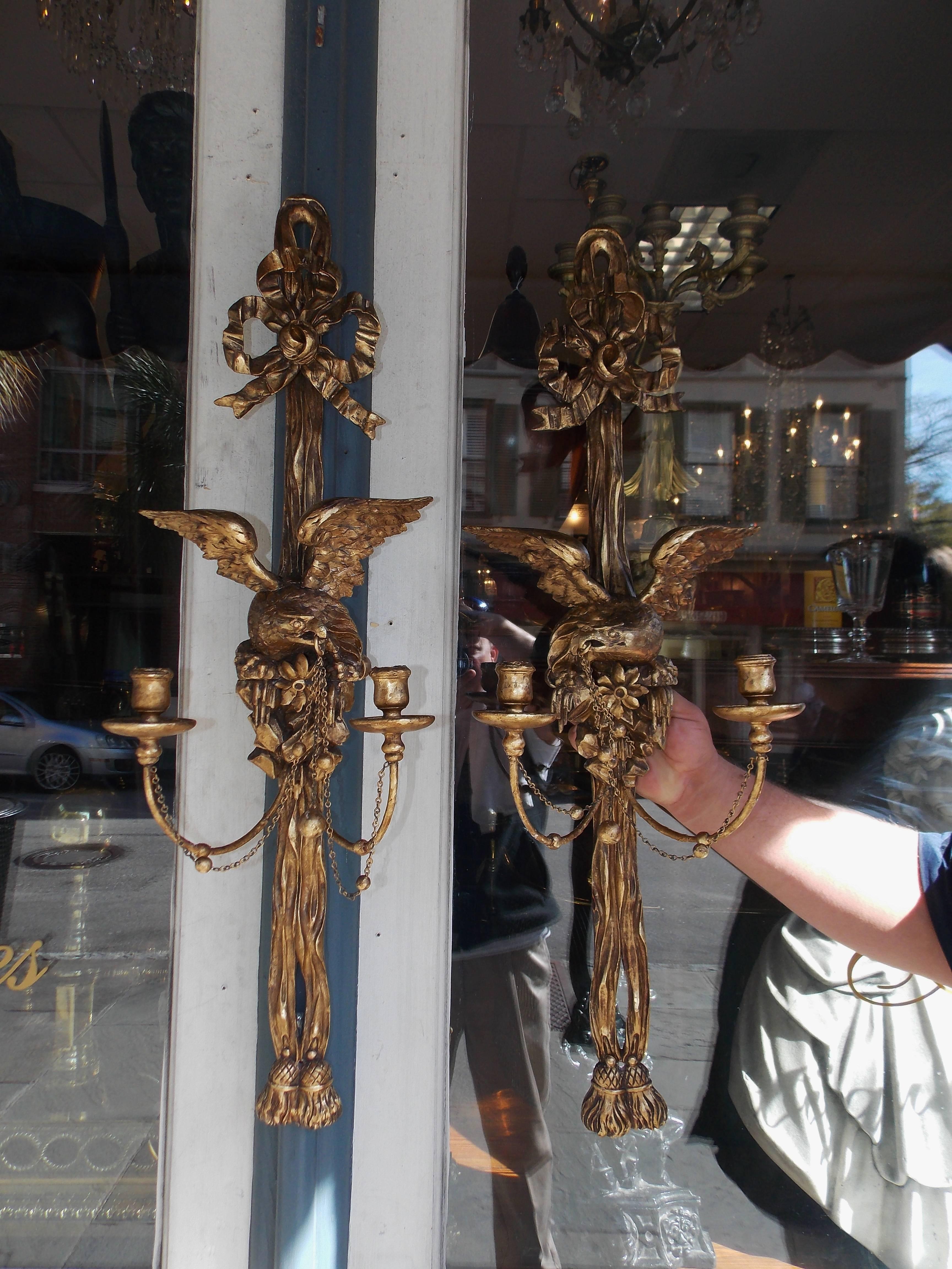 Pair of American gilt carved wood two-arm wall sconces with perched facing eagles to flee on rocky plinths, attached chain with circular spheres, flanking ribbon motif and terminating with a double lower tassel. Early 19th century. Sconces are