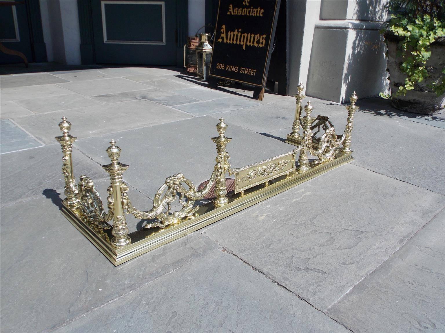 French brass fire fender with flanking urn finial columns, laurel wreaths, connecting swags, ribbon motifs, a centered floral rectangular panel, and resting on a molded step back base, Early 19th Century.