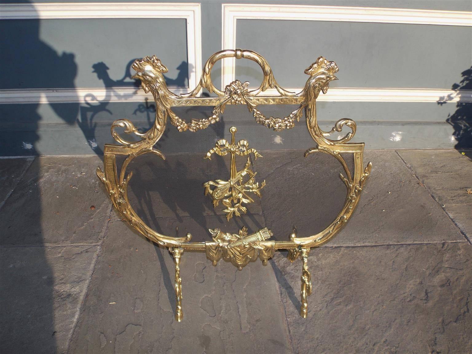 Parafuoco francese in ottone con teste di gallo affiancate, maniglia centrale a fogliame, festoni floreali, nastro decorativo circondato da strumenti musicali, torce inferiori appoggiate su nappe, terminante su quattro gambe a volute d'acanto con