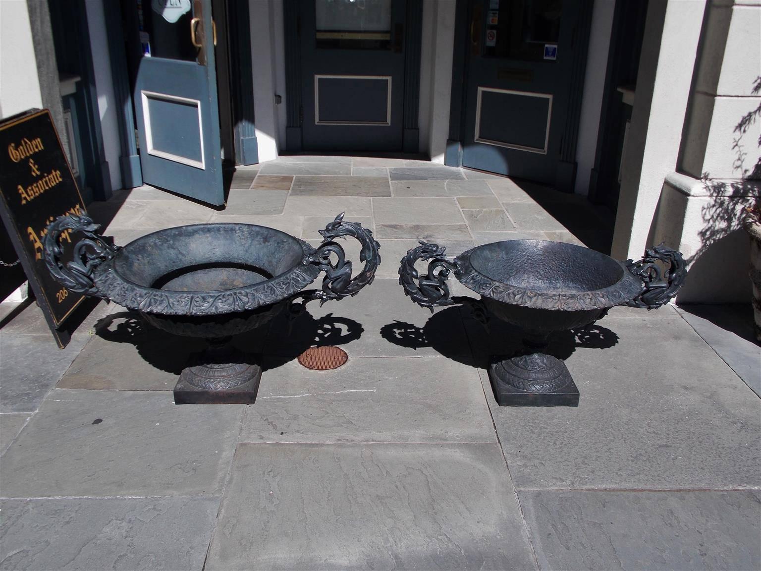 Pair of American cast iron garden planters with floral molded edges, scrolled dragon handles, and terminating on raised decorative floral circular and squared plinths, Early 19th Century. 

Measures: Large urn 48