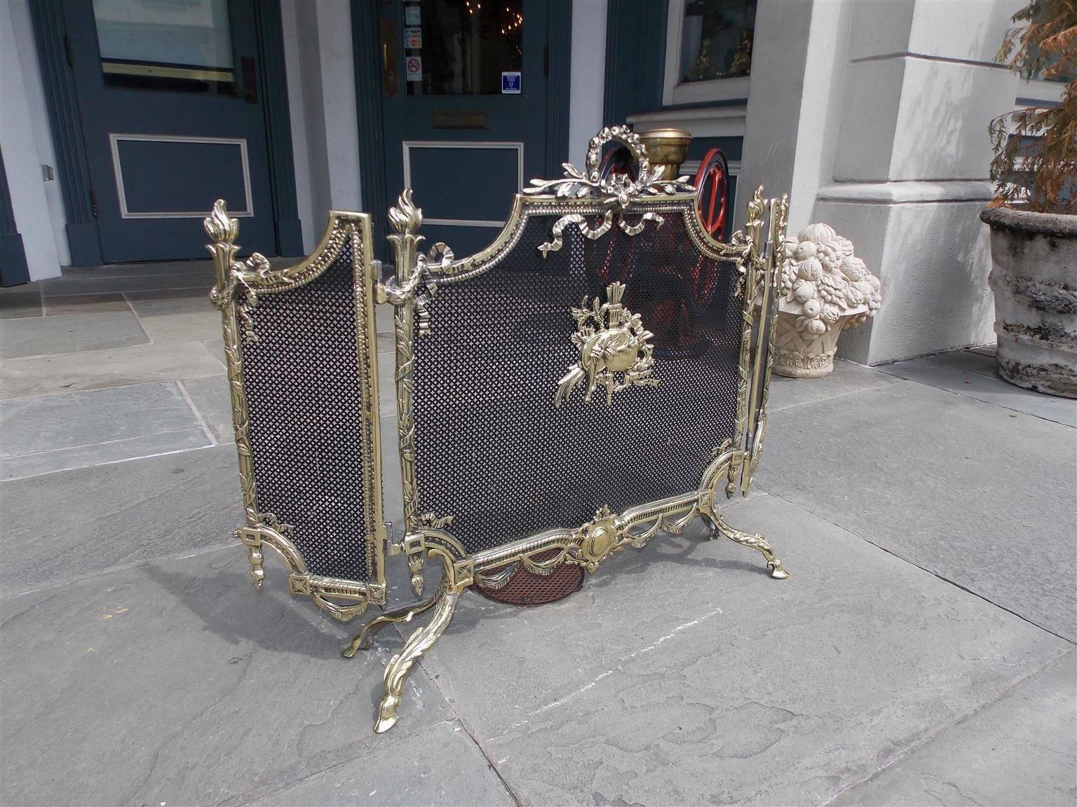French brass folding fire screen with a decorative laurel wreath ribbon handle, flanking flame corner finials, centered arrow medallion, lower ornate medallion with swag motif, and terminating on acanthus scrolled hoof feet, Early 19th Century.