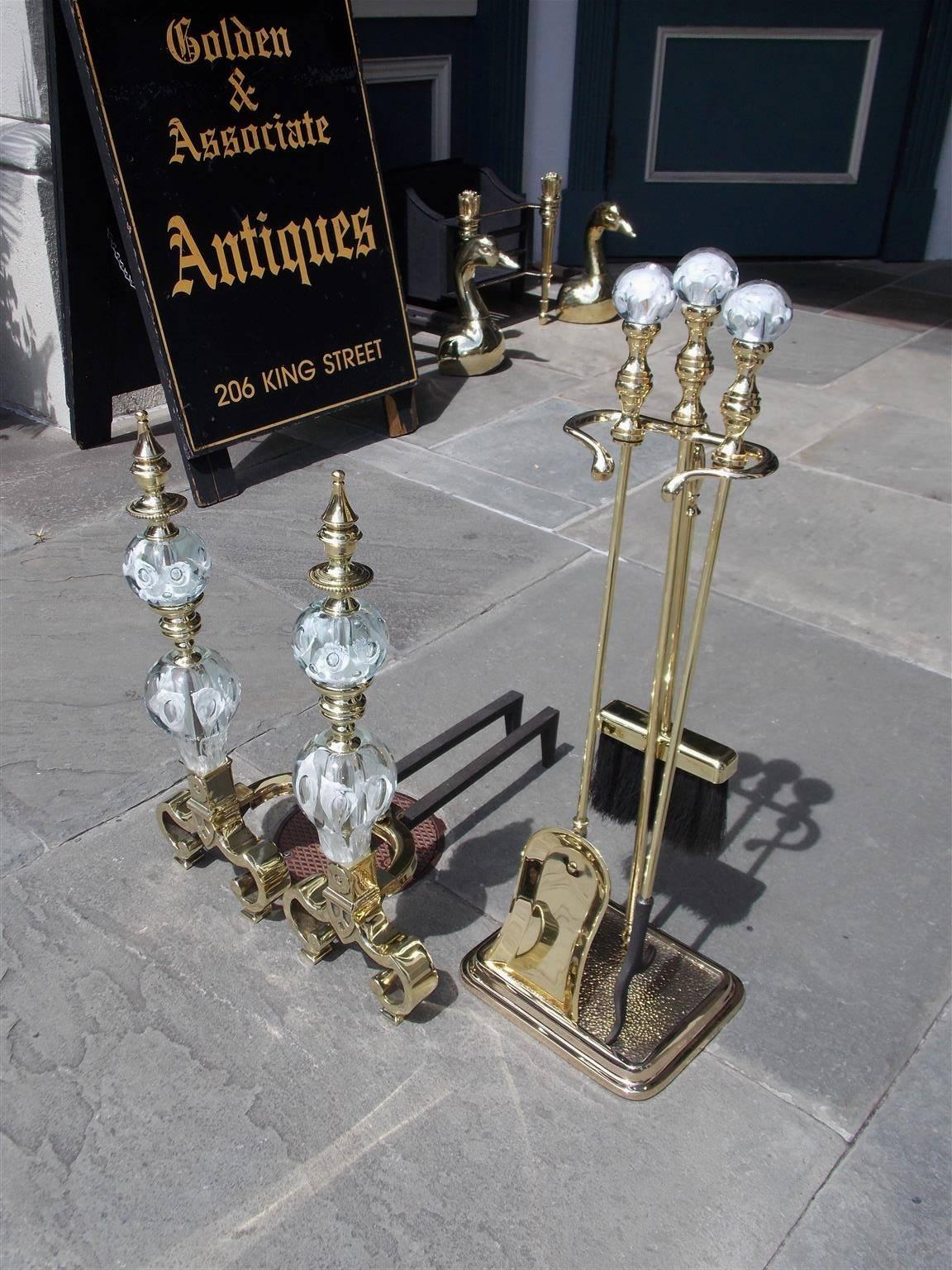 Set of American brass St. Clair andirons with matching tools on stand. The clear globes contain controlled bubbles and feature groupings of stylized white flowers. Andirons consist of flanking finial tops, crystal sphere balls, turned bulbous