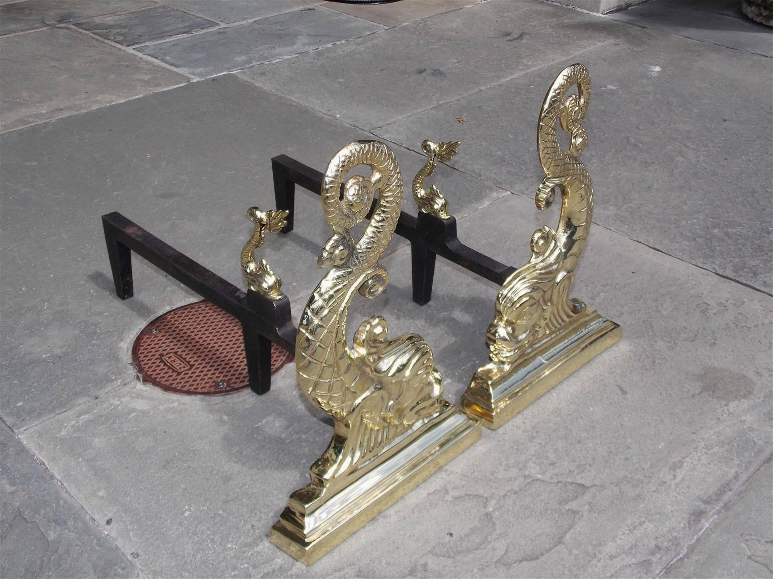 Pair of American brass decorative dolphin fire place andirons with flanking heads, scrolled intertwined engraved tails, matching miniature log stops , original iron legs, and resting on step back molded edge plinths, Mid-19th century.