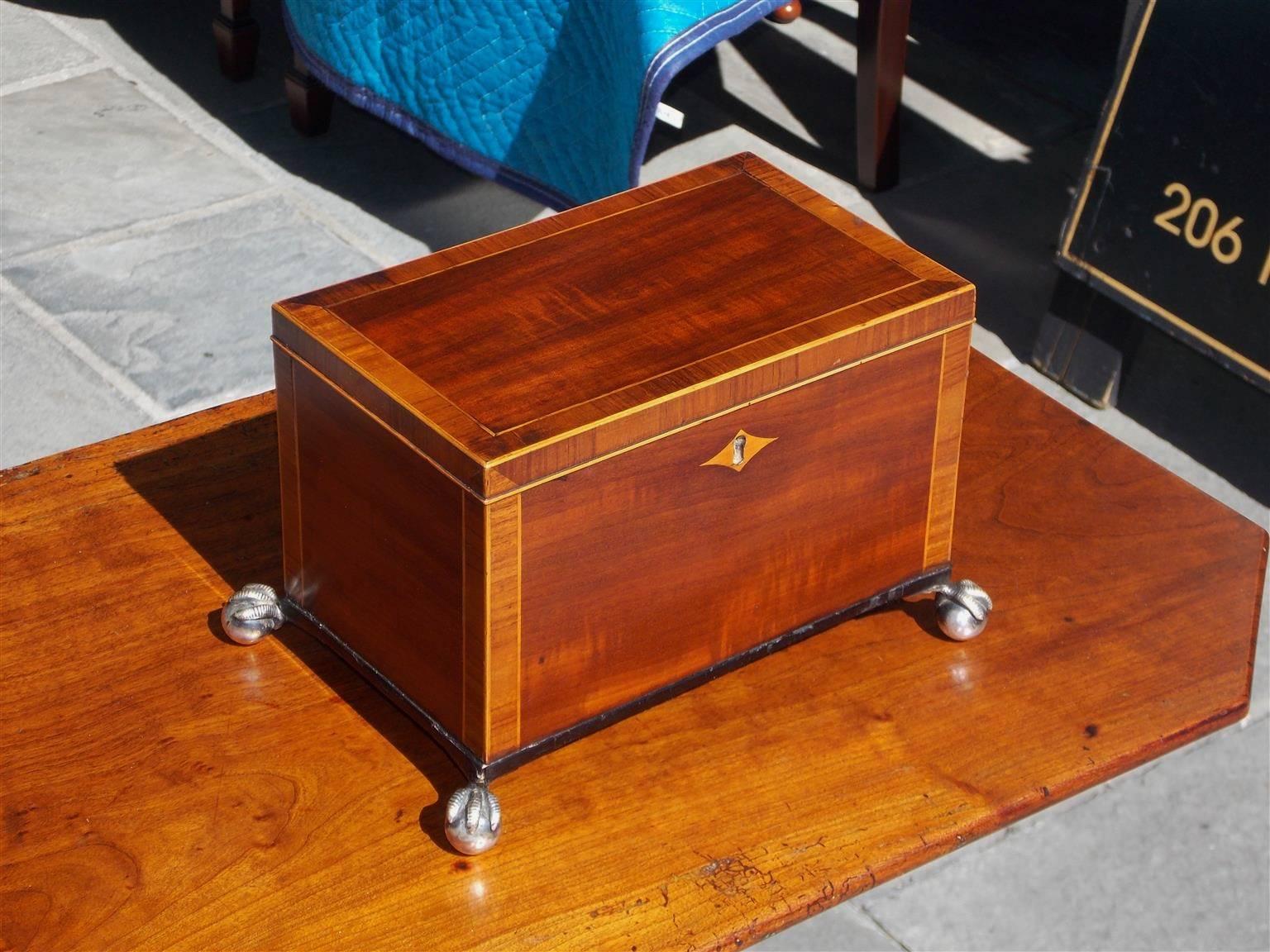 English Regency mahogany tea caddy cross banded in tulip wood with satinwood string inlays, centered diamond escutcheon, hinged lid with compartmentalized lined interior, and terminating on ebonized molded base with silver over brass ball and claw