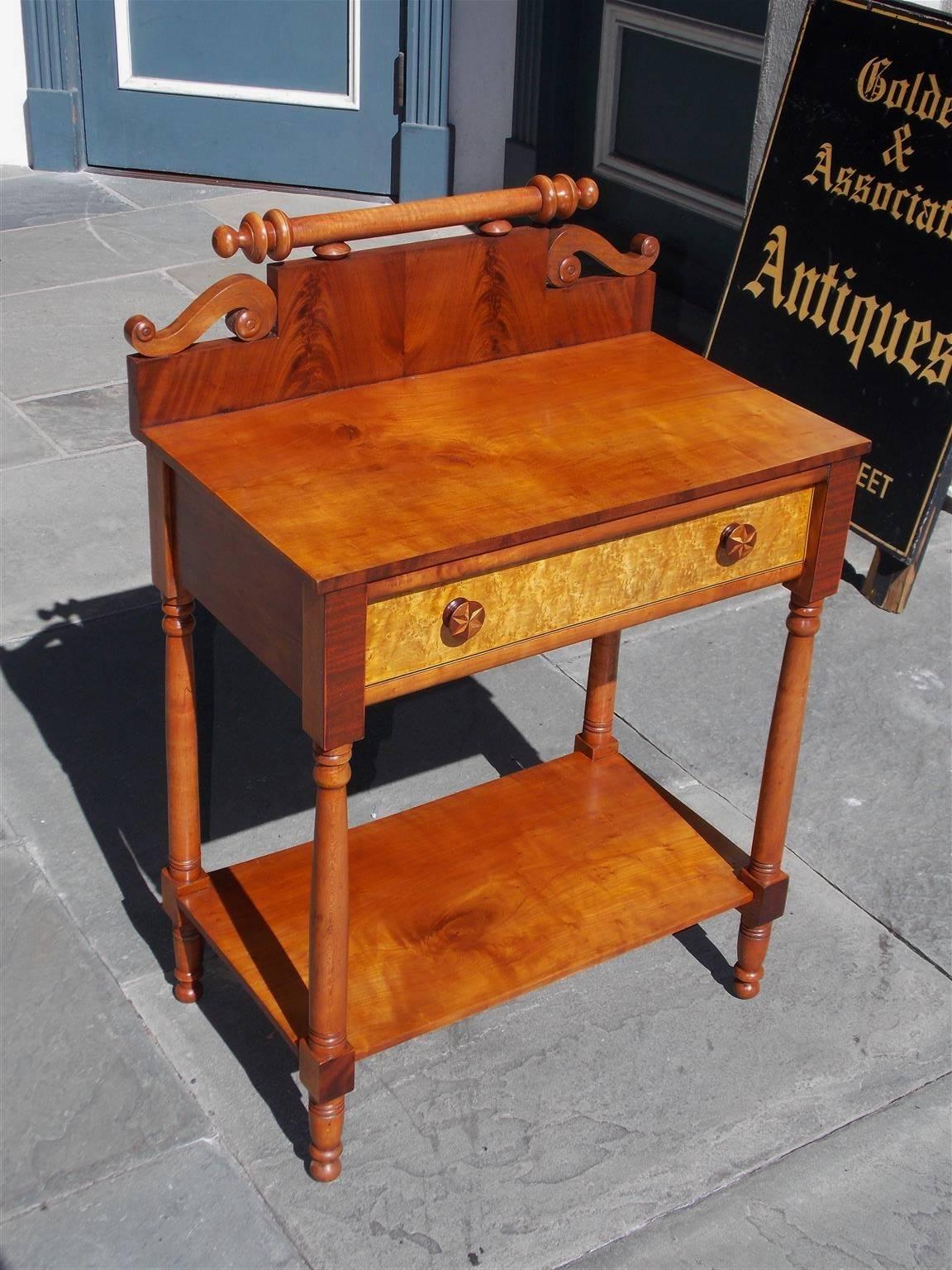 American Sheraton mahogany and cherry one drawer side table with a carved scrolled book matched back splash, inlaid bird's-eye drawer front, original star inlaid wooden knobs, lower case shelf, and terminating on turned bulbous ringed legs, Early