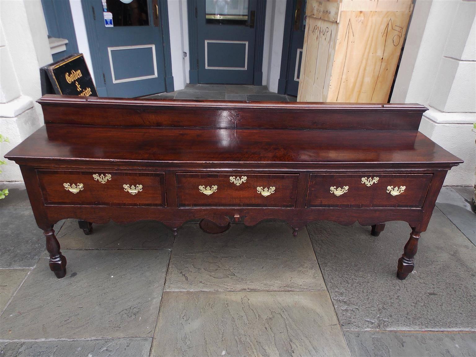 English oak console with a molded edge floral inlaid back splash, three fitted cross banded drawers with the original brasses, carved scalloped skirt with flanking centered finials, and terminating on the original turned bulbous squared legs, Late