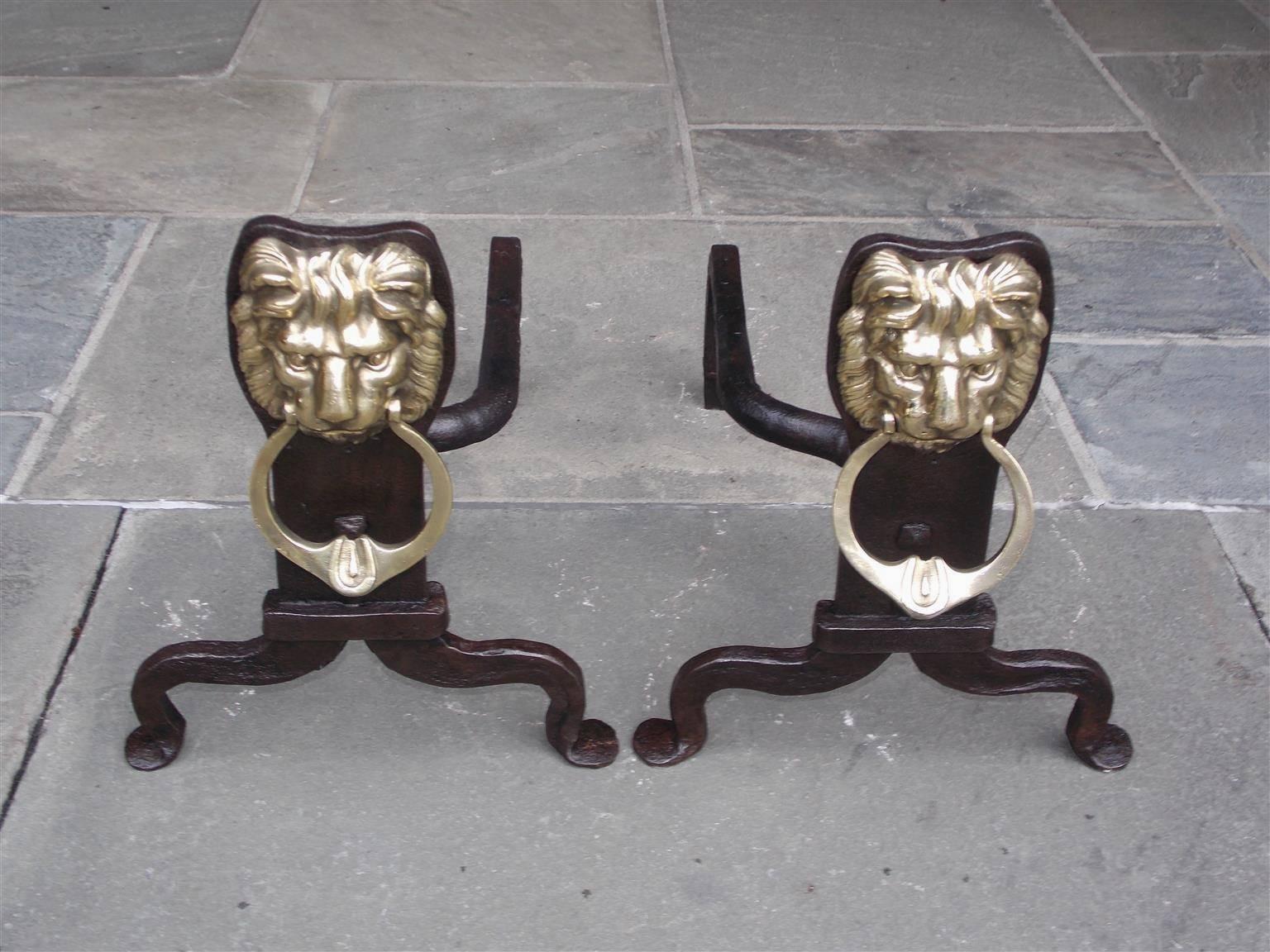 Pair of English Lion face brass and wrought iron andirons with flanking oval rings mounted in the corners of mouth, squared centered plinths with rare reverse side spit hooks for poker and terminating on the original scrolled penny feet, Late 18th