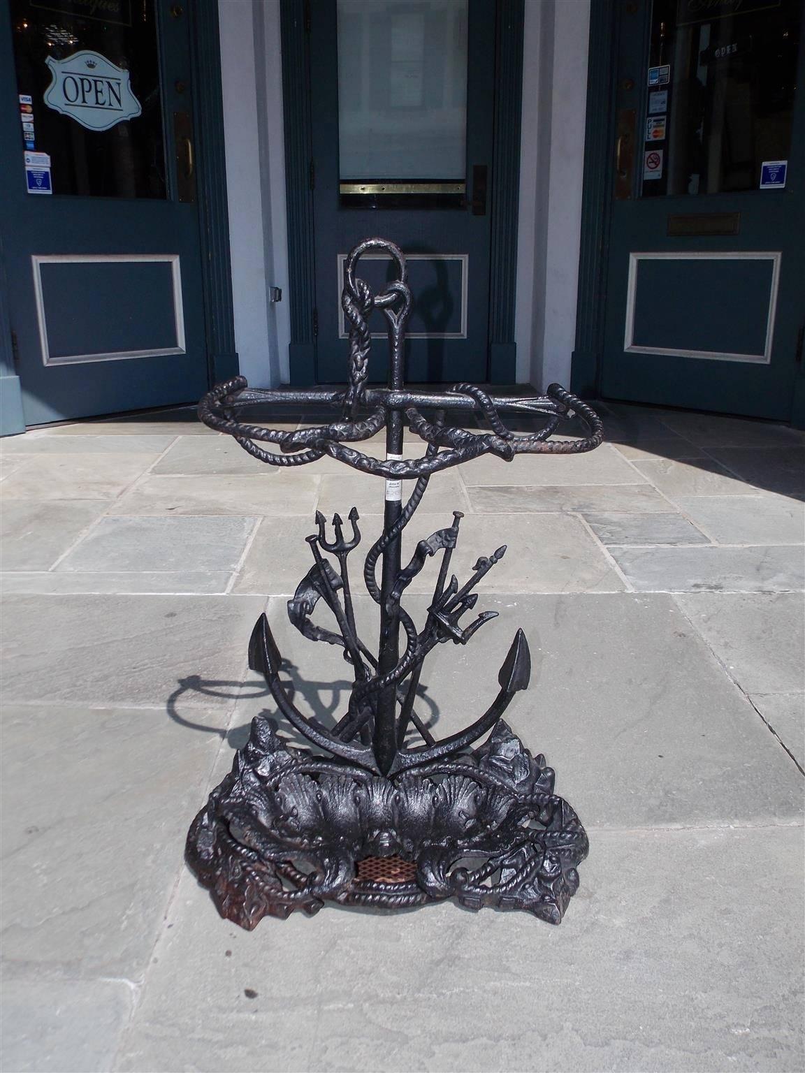 American cast iron nautical umbrella stand with centered anchor ring attached and intertwined with rope, two smaller tridents with spears, and resting on a stylized rocky base with a removable shell and dolphin motif pan, Mid-19th century.