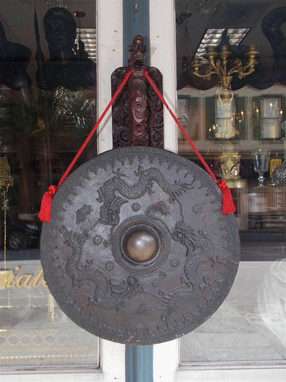 Chinese king wood circular temple gong with a decorative carved wall-mounted dragon bracket supporting a braided silk rope bronze gong with decorative dragons, floral medallions, and Koi fish. Gong has the original mallet. 19th Century. Wall-mounted