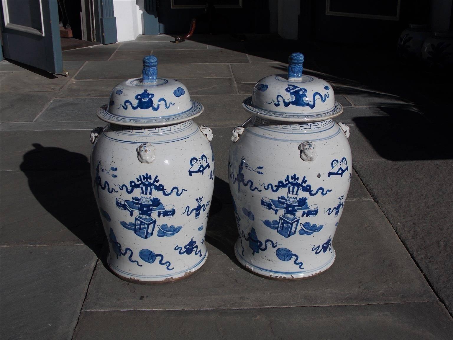 Pair of Chinese porcelain glazed blue and white temple jars with flaking foo dogs finials, decorative painted borders, surrounding figural faces, and centered painted vases with floral motif.  Lids are removable and were used to store perishable