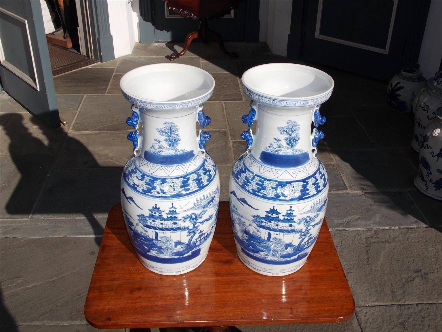 Pair of Chinese porcelain glazed blue and white temple jars with hand-painted Greek key borders, flanking foo dog side handles, and decorative hand-painted pagoda, mountain, and landscape scenes. 20th century.