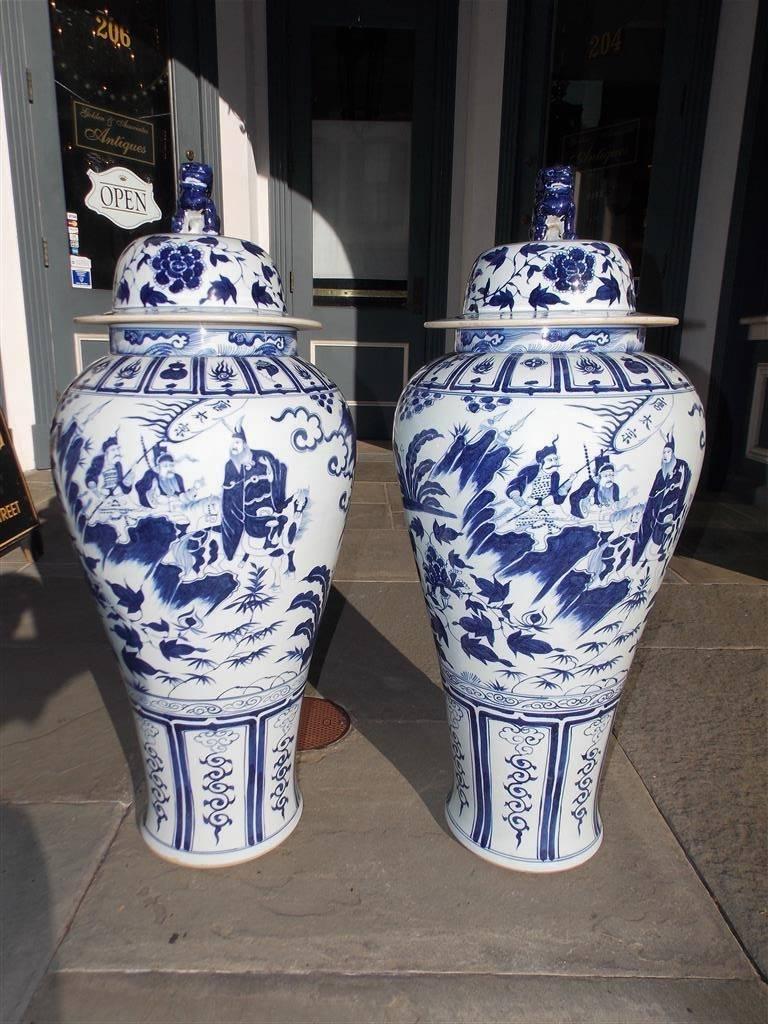 Pair of monumental Chinese porcelain glazed vibrant painted blue and white temple urns with removable foo dog finial lids, warriors in battle mounted on horses, landscapes with cascading streams, and resting on decorative floral tapered bases, 20th
