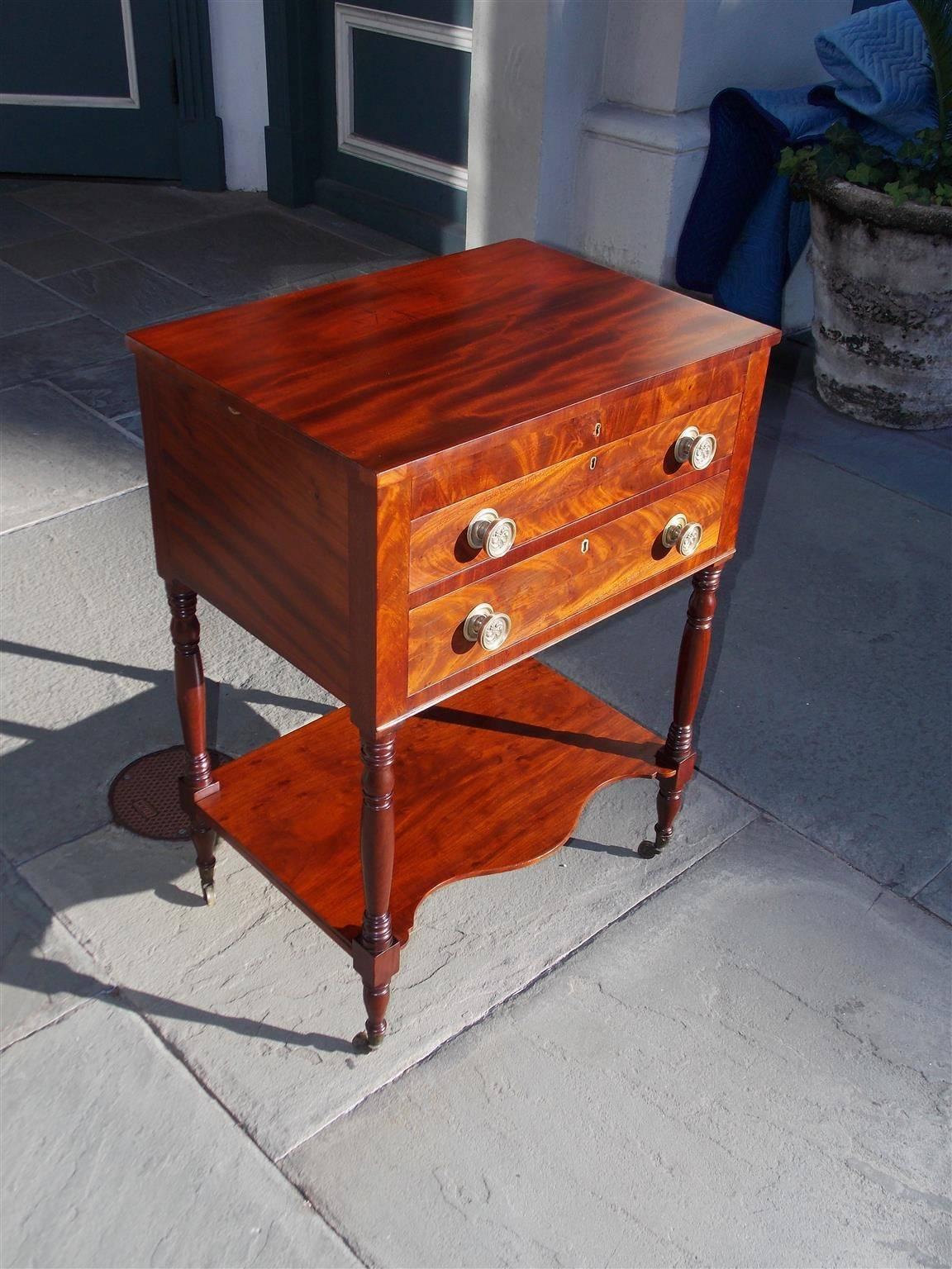 American Sheraton mahogany two-drawer work table with a hinged top revealing a fitted interior compartmentalized desk , original pinwheel brasses, serpentine lower shelf, and terminating on turned bulbous ringed legs with the original brass casters,