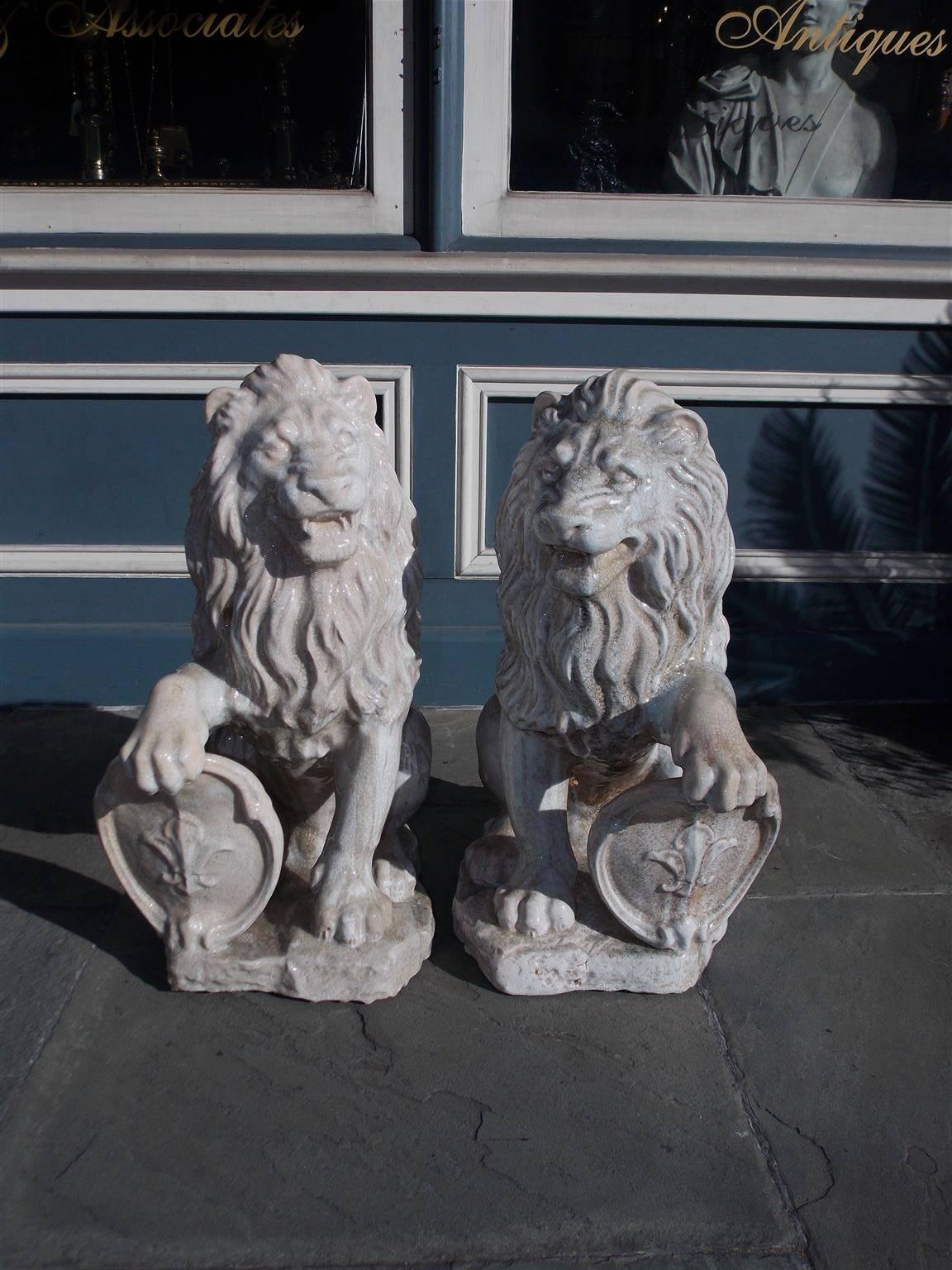 Pair of French painted and glazed terracotta lions grasping oval Fleur De Lis plaques while resting on rectangular plinths. Mid-19th century.