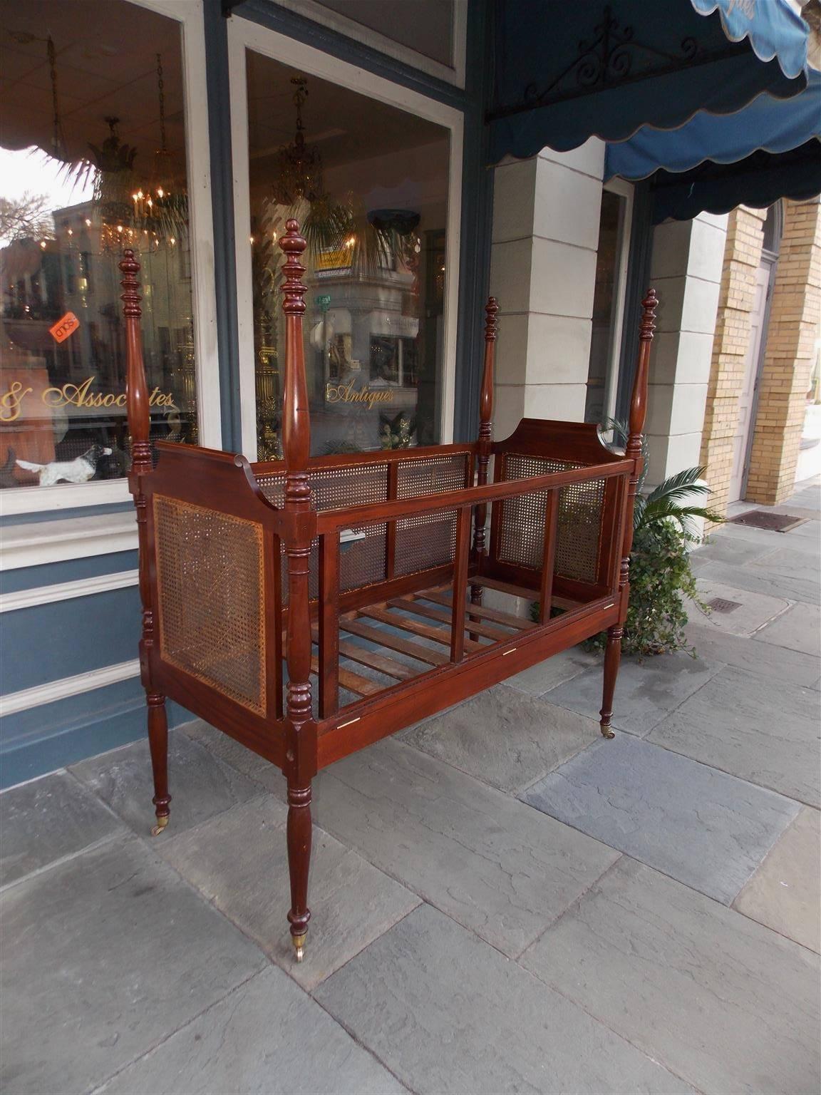 Charleston mahogany crib with flanking scrolled cane headboards, four urn finial bulbous turned ringed post, hinged finger locked swing down side panels, original side rails with supporting poplar slats, and terminating on the original brass