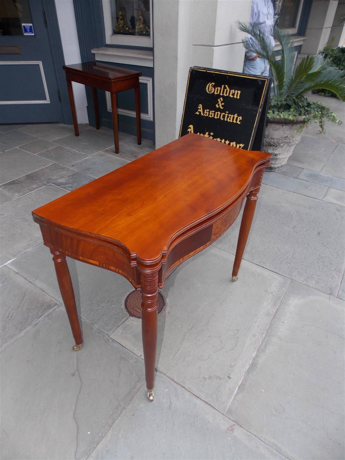 American Sheraton cherry serpentine game table with a carved reeded edge, outset cut corners, centered lace wood plate surrounded by tulip wood inlay, and terminating on the original turned ringed legs with the original brass casters. Minor warping