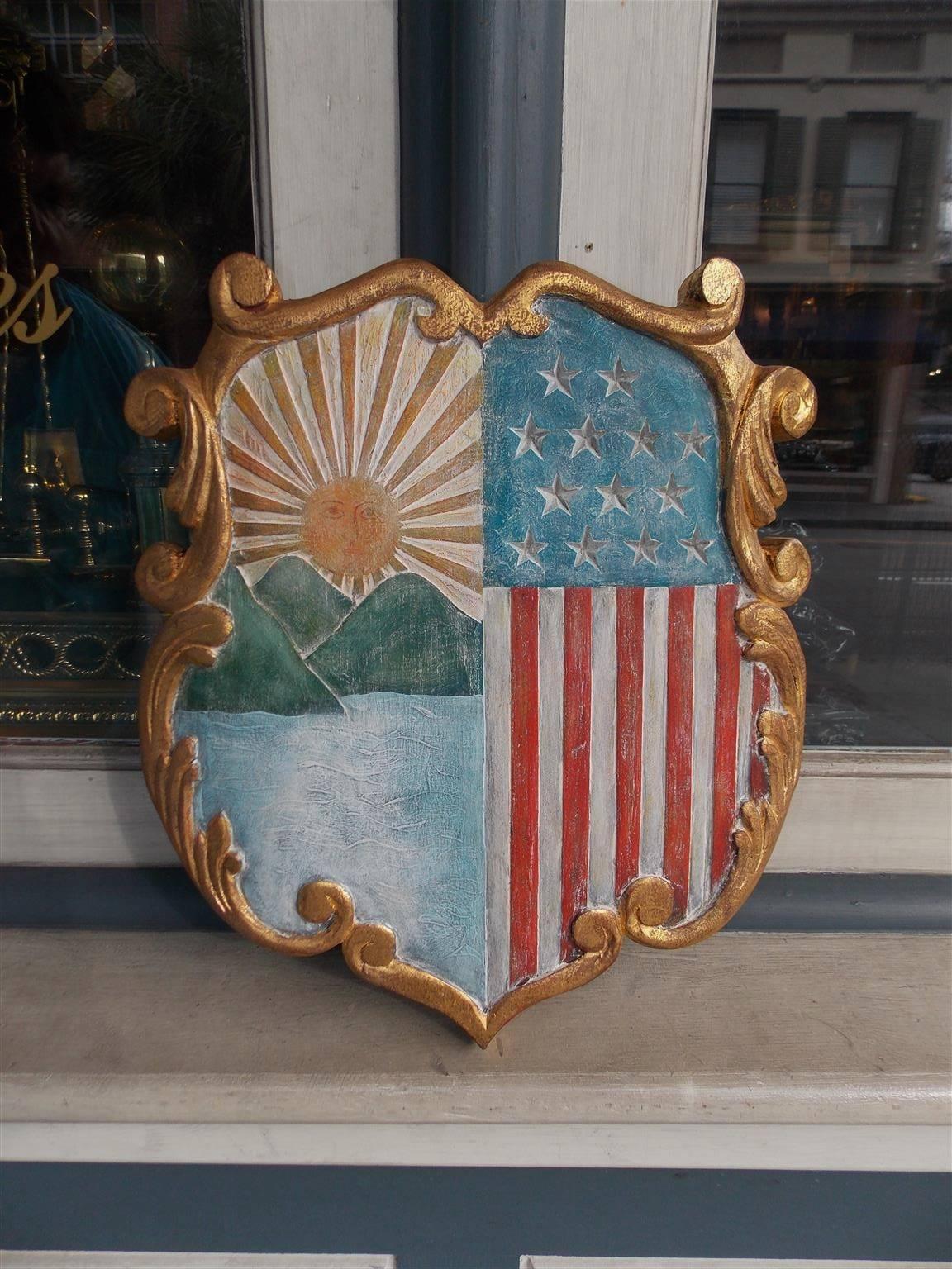 American hand-carved wood gilded and painted patriotic shield depicting the American flag , Sun, Mountains, Ocean with an artistic scrolled acanthus gilt border, Early 20th Century.
