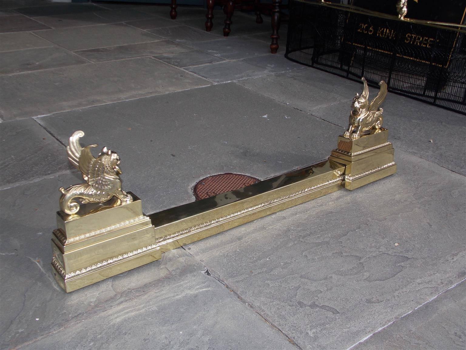 French brass Chenet with flanking winged griffons, decorative hand chased egg and dart motif, lambs tongue motif, and terminating on rectangular plinths, Early 19th century.