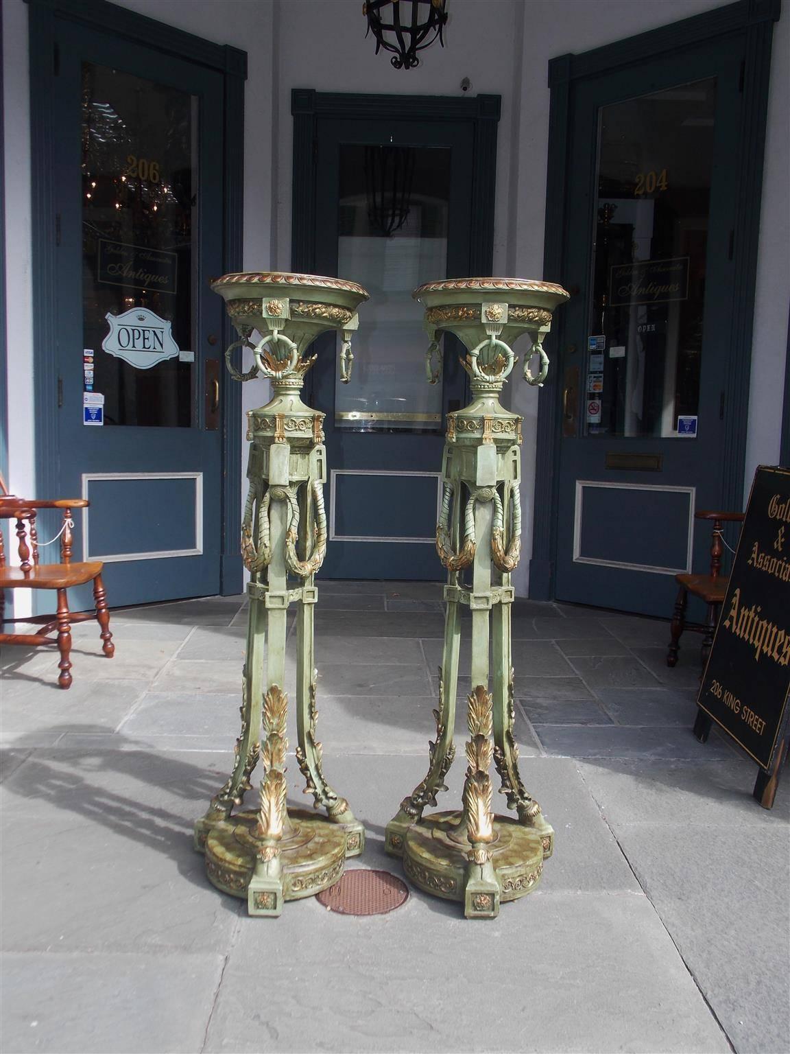 Pair of Italian painted and gilt torchieres with circular egg & dart medallion tops, floral decorative rings, intertwining floral tassels and terminating on acanthus tripod hoof feet resting on circular foliage bases with centered flamed spheres,
