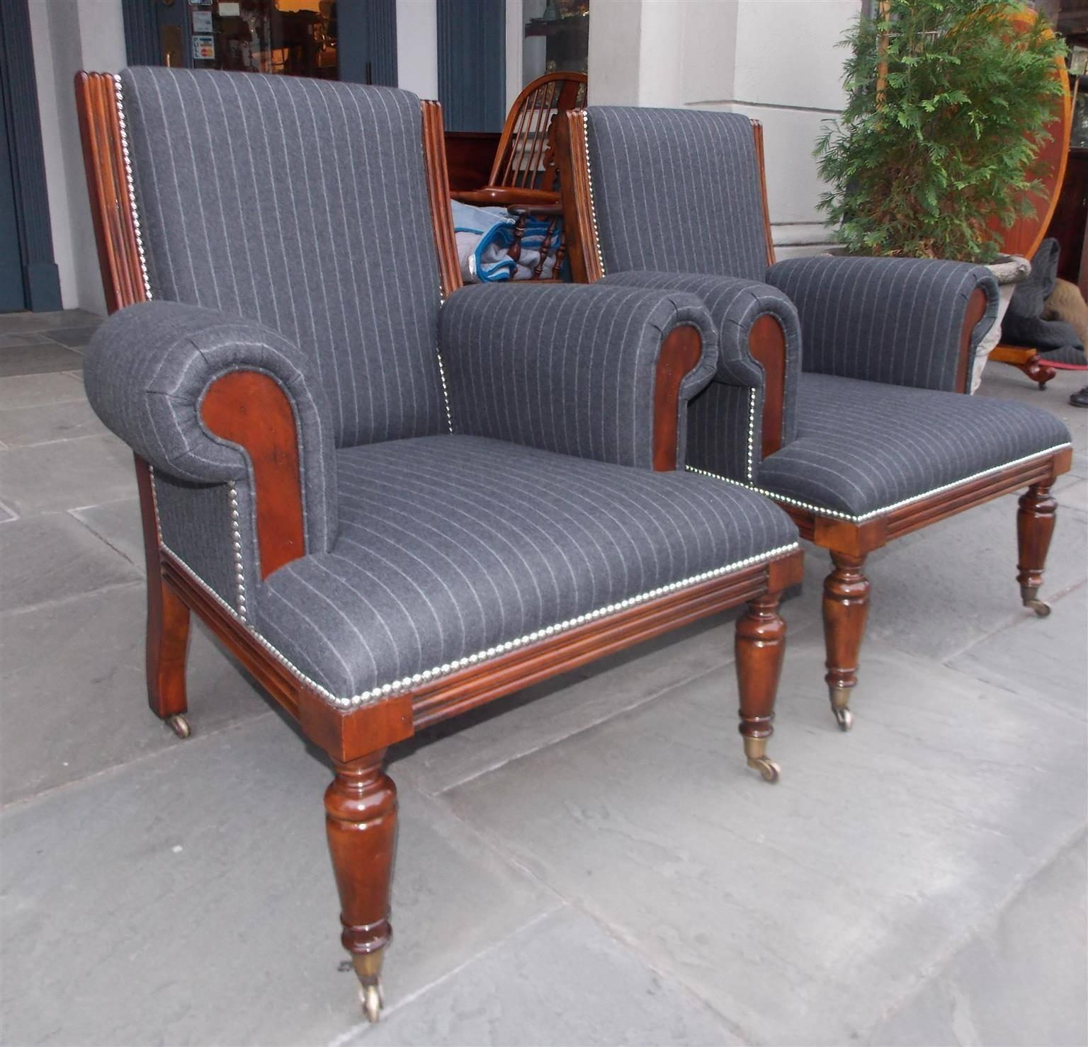 Pair of American mahogany club chairs with carved reeded backs, scrolled arms, nickel tacking, and terminating on turned bulbous legs with original brass casters.  20th Century.  Chairs are upholstered in a pin strip wool fabric.  