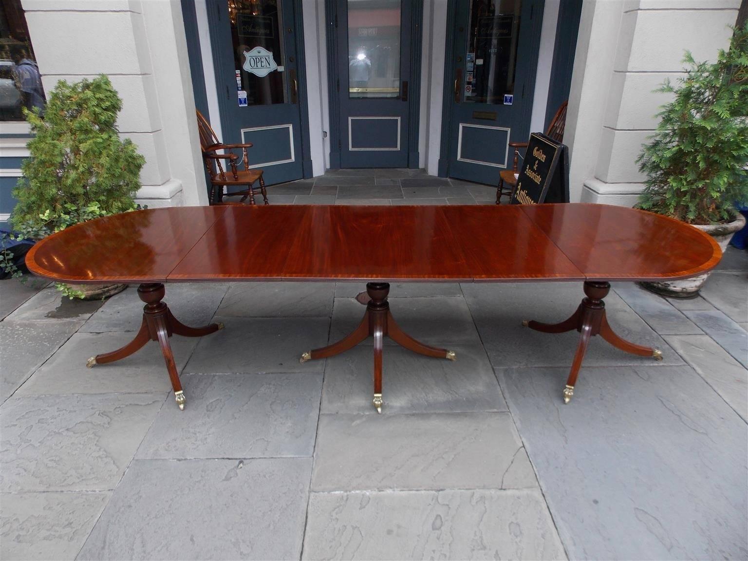 English mahogany triple pedestal dining room table with satinwood Inlaid banding, two removable leaves, and terminating on turned bulbous saber legs with original brass paw casters, early 19th Century. Dining table is 124.25