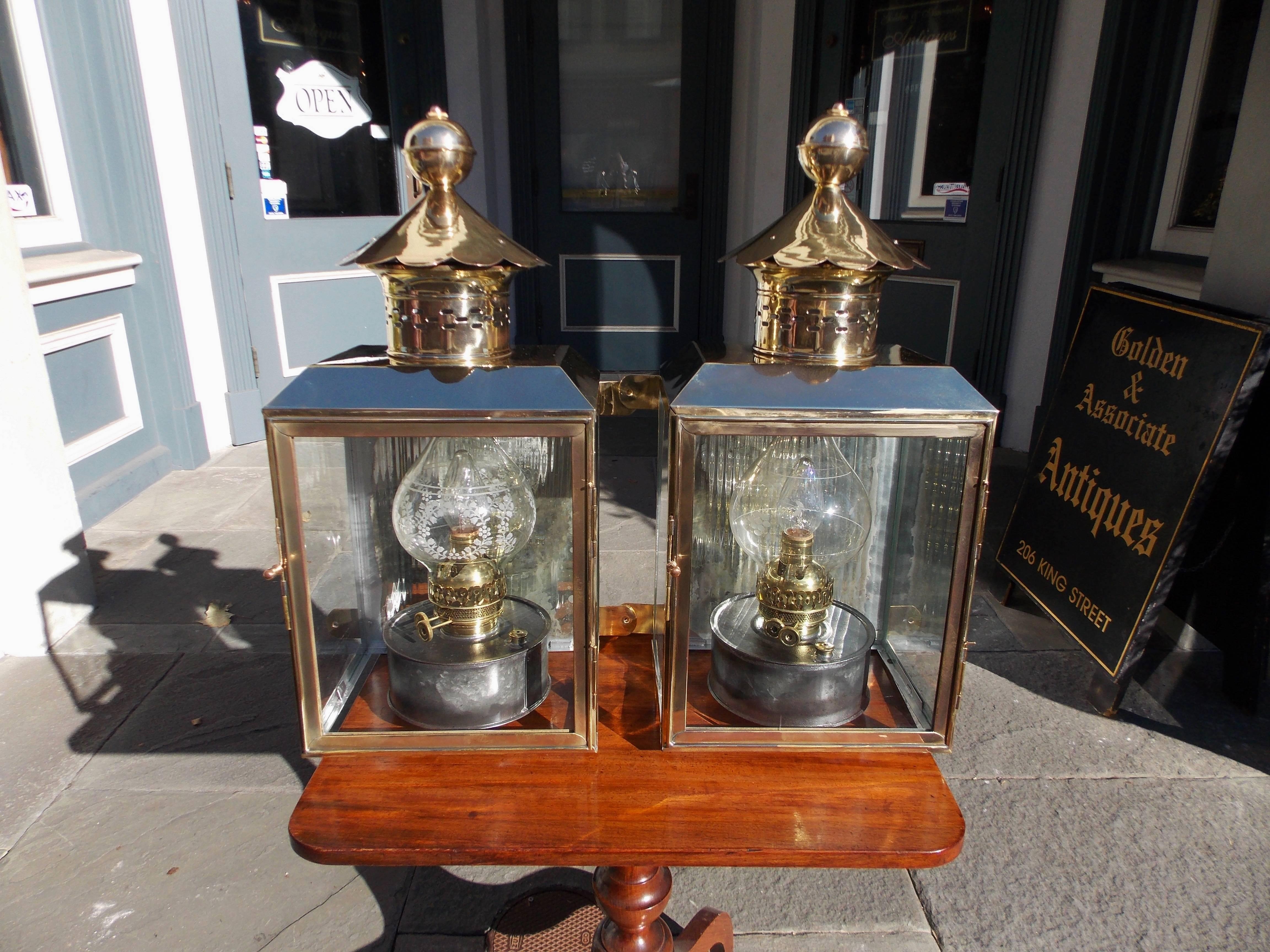 Pair of American brass and polished steel wall lanterns with vented finial domes, glass case panels with a hinged door, side mounting brackets, and the original polished steel kerosene burners with acid etched shades. Lanterns have been converted to