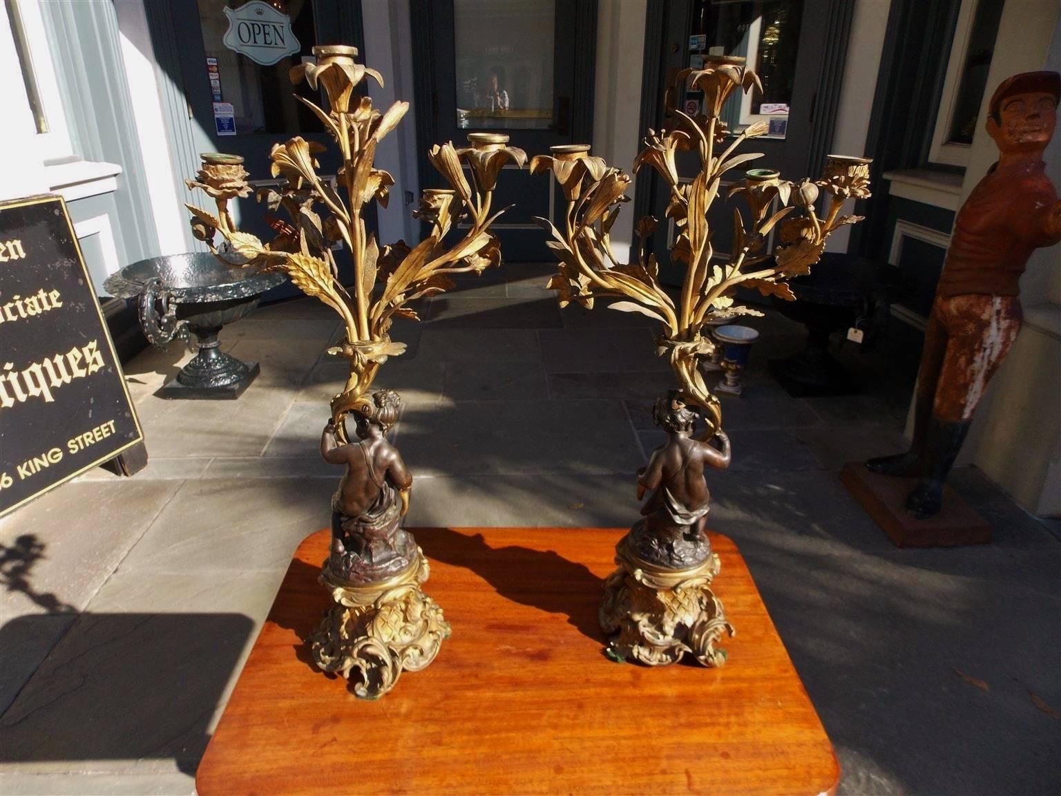 Pair of French ormolu and bronze five-arm candelabras with flanking cherubs, candle bobeches, and resting on circular plinths with basket weave, floral & shell motif, Early 19th century. Candle powered and can be electrified if desired, Early 19th