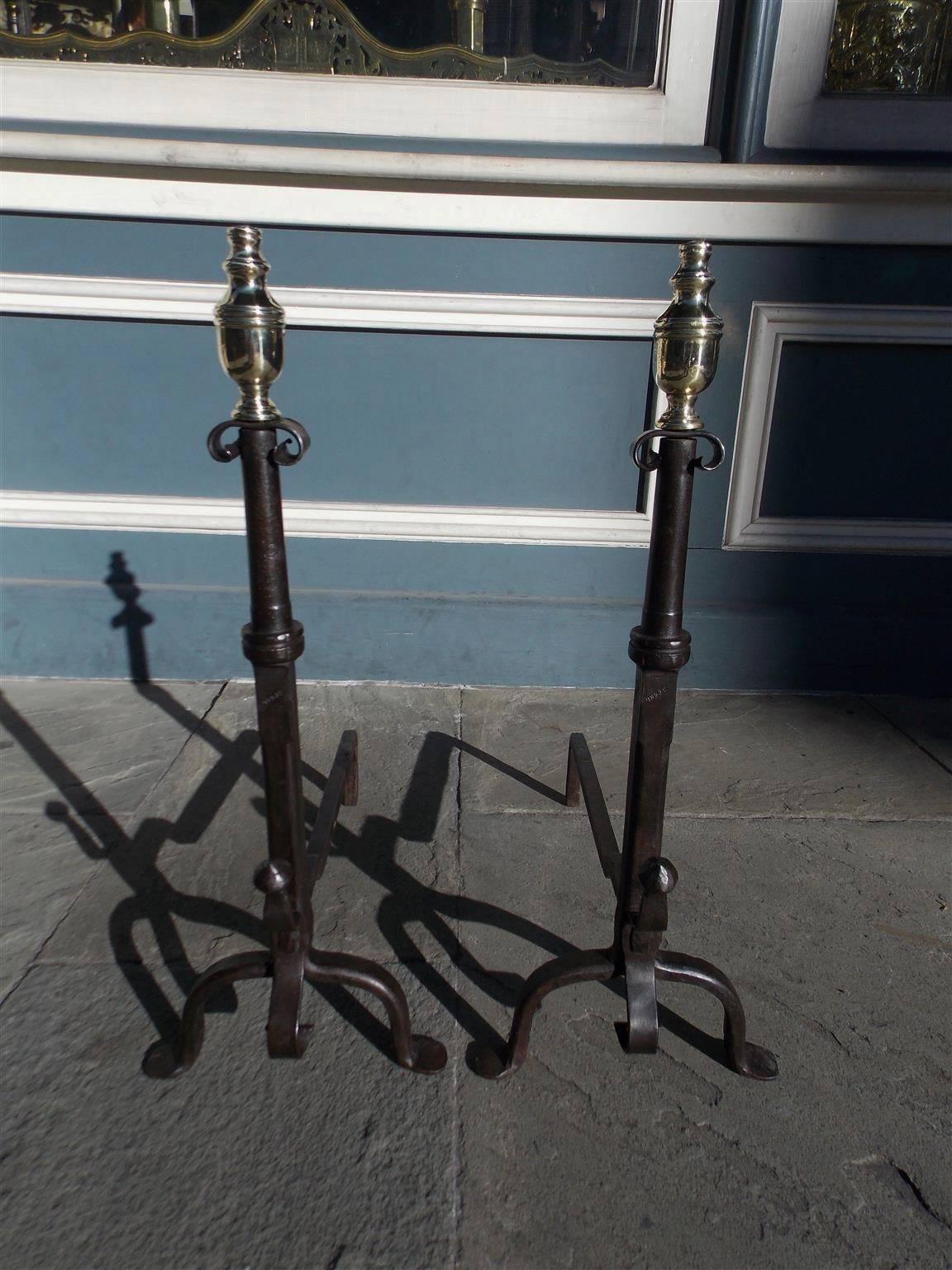 American wrought iron and brass andirons with urn top finials, scrolled circular squared plinths, spit hooks, and terminating on penny feet.  Late 18th Century.  
Center plinths were originally axles on a horse drawn carriage. 