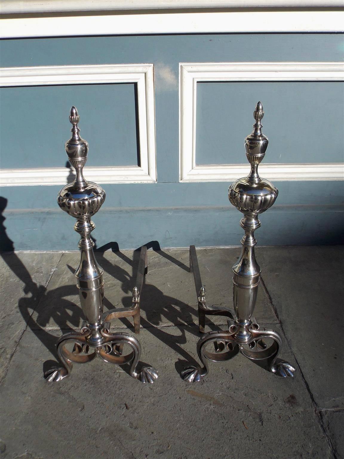 English nickel silver and wrought iron andirons with urn fluted acorn finial tops, bulbous turned ringed plinths, matching acorn finial log stops, decorative scrolled legs, and terminating on stylized lions paw feet. Late 18th Century