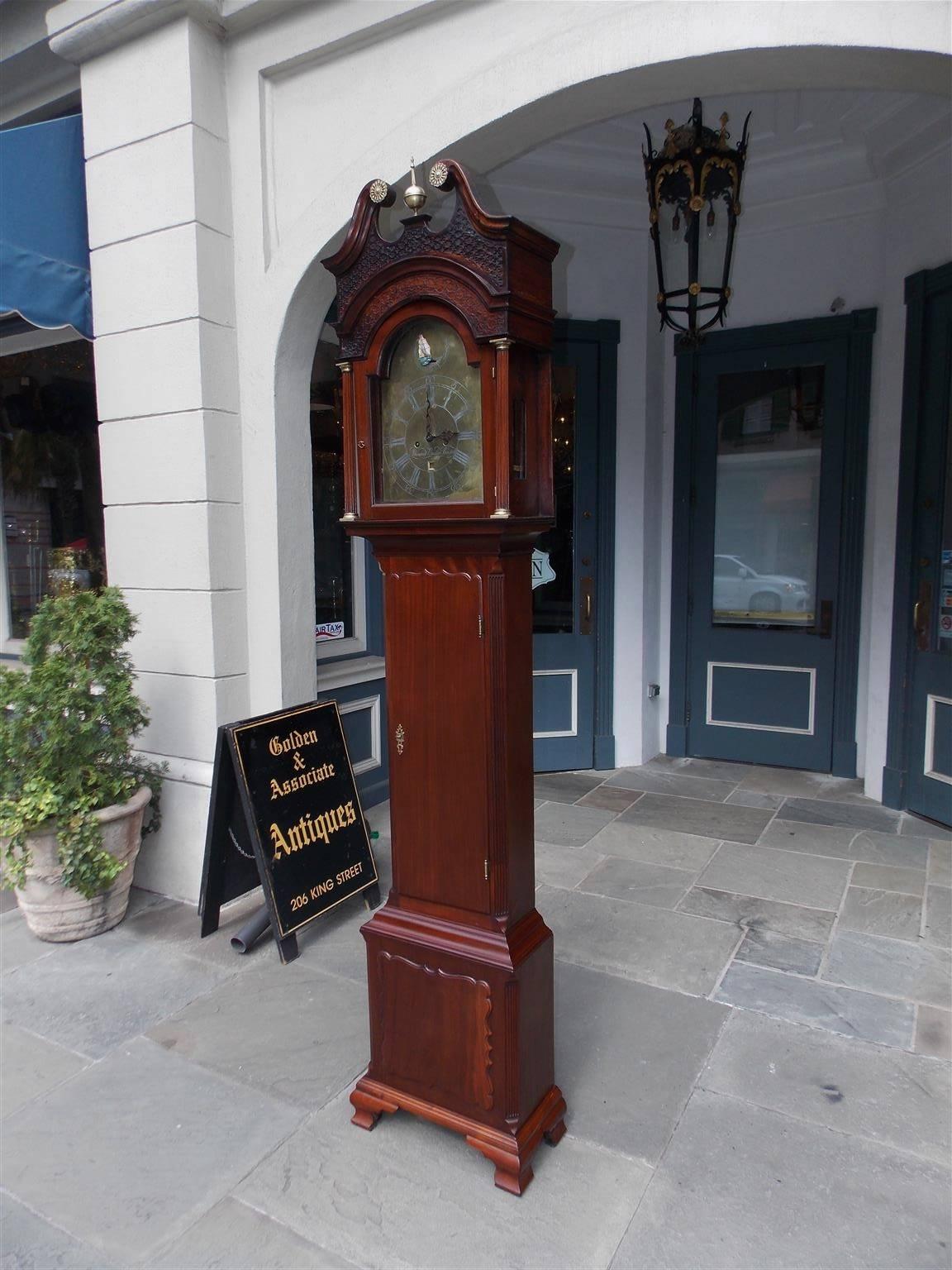 English Chippendale mahogany broken pediment tall case clock with centered brass urn finial, brass floral filigree medallions, intricately carved fret work, fluted columns with brass mounts, brass face with engraved numerals, rocking ship, carved