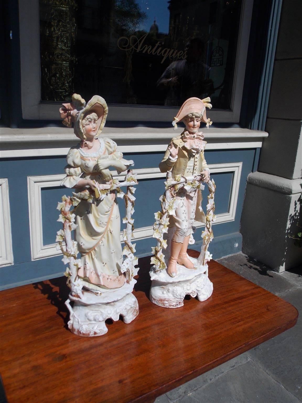 Pair of French Bisque figurines standing and greeting people in formal dress attire leaning against wooden post with decorative ivy motif and resting on oval floral plinths, Late 19th Century.