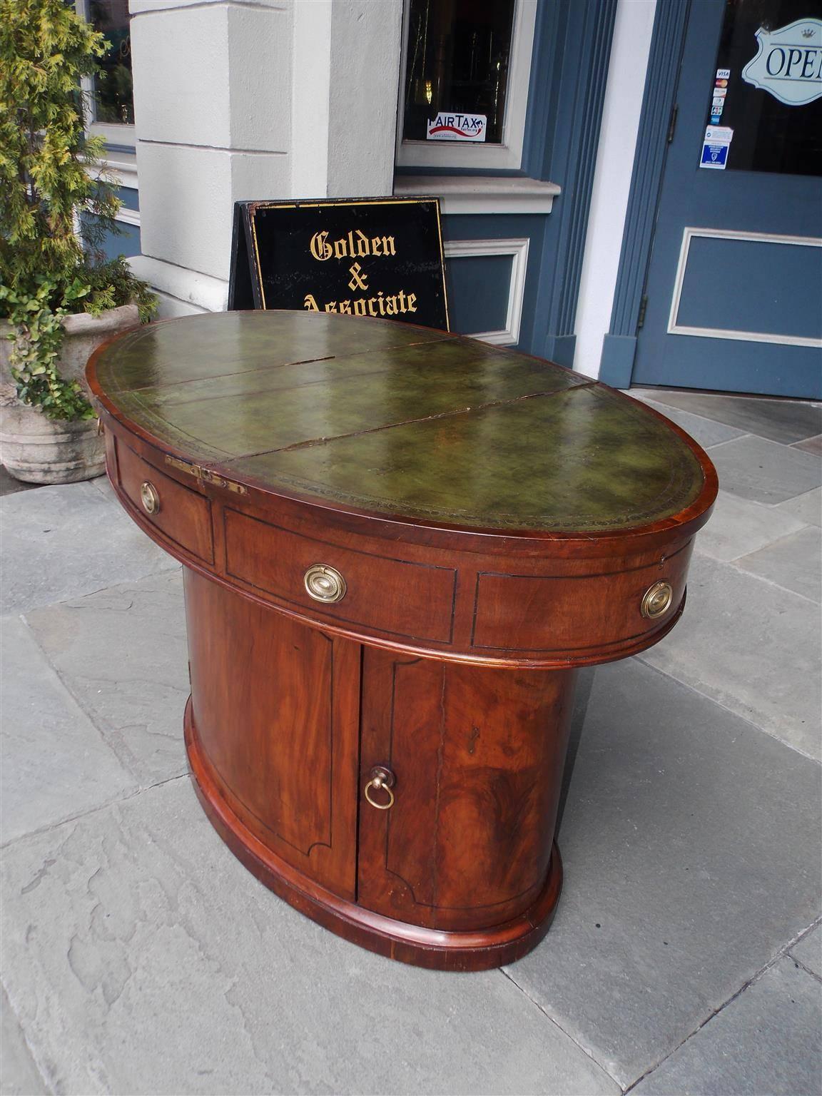 English mahogany oval sea captains desk with hinged leather top concealing two upper hidden compartments, single front drawer, original brasses, ebony string inlay, two hinged lower cabinets with interior shelving, and resting on a carved molded