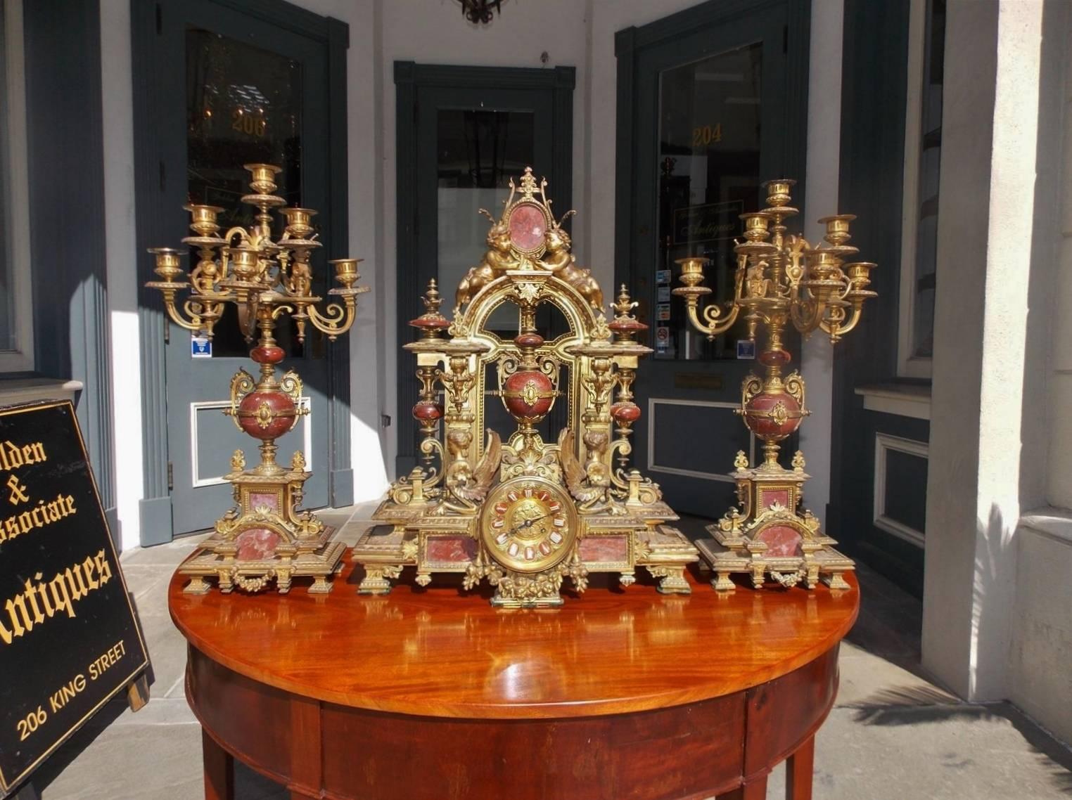 French gilt bronze and marble three piece mantel clock flanked by matching candelabras. Center clock has cherub, griffon, and floral fruit motif. Clock is in working condition.