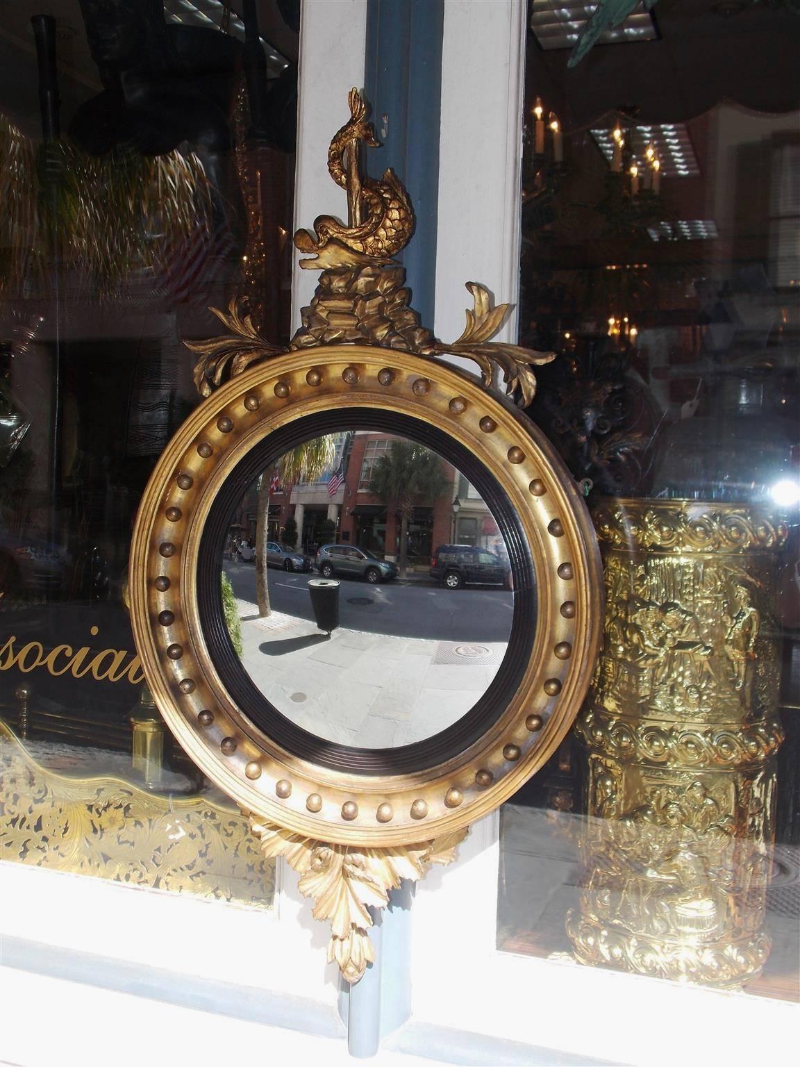 American Federal gilt carved wood convex mirror with a perched intertwined dolphin on a rocky ledge, ebonized reeded banding, circular ball motif, and adorned with foliage pendants. Early 19th Century