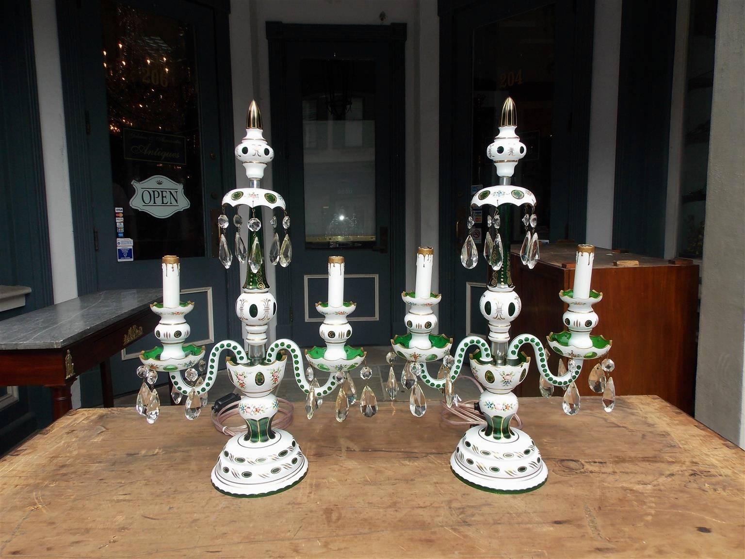 Pair of Bohemian emerald green glass table lamps with flanking gilt spheres, inverted bobeches with hanging crystal prisms, bulbous centered floral columns, two tiered bobeche scrolled arms with hanging crystal prisms, and terminating on a