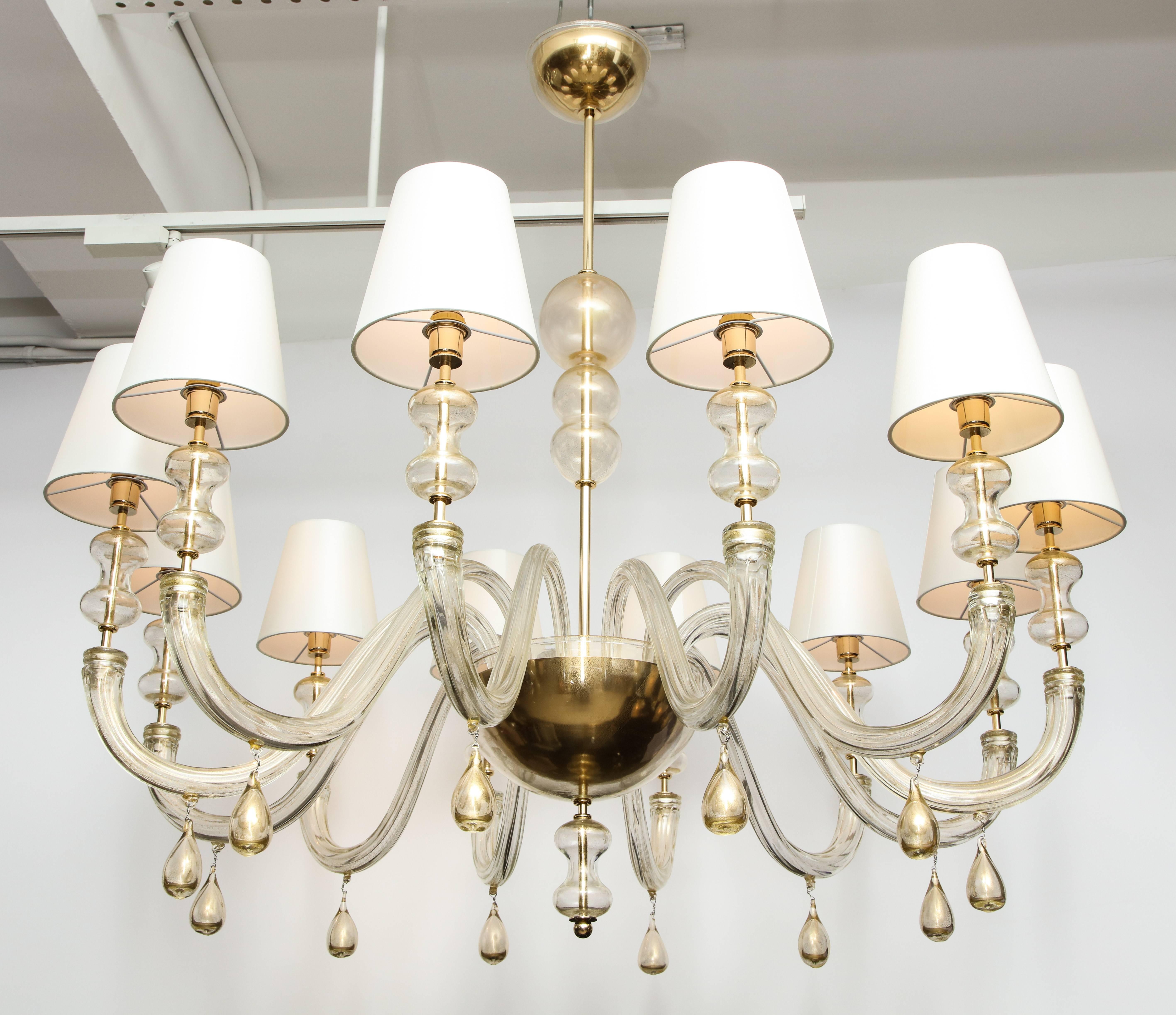 Classic but with a modern twist. This elegant Italian modernist chandelier in Murano clear glass with 23-karat gold flecks is comprised of 12 arms, each glass arm is adorned with a glass drop and a silk ivory lampshade the clear glass is infused
