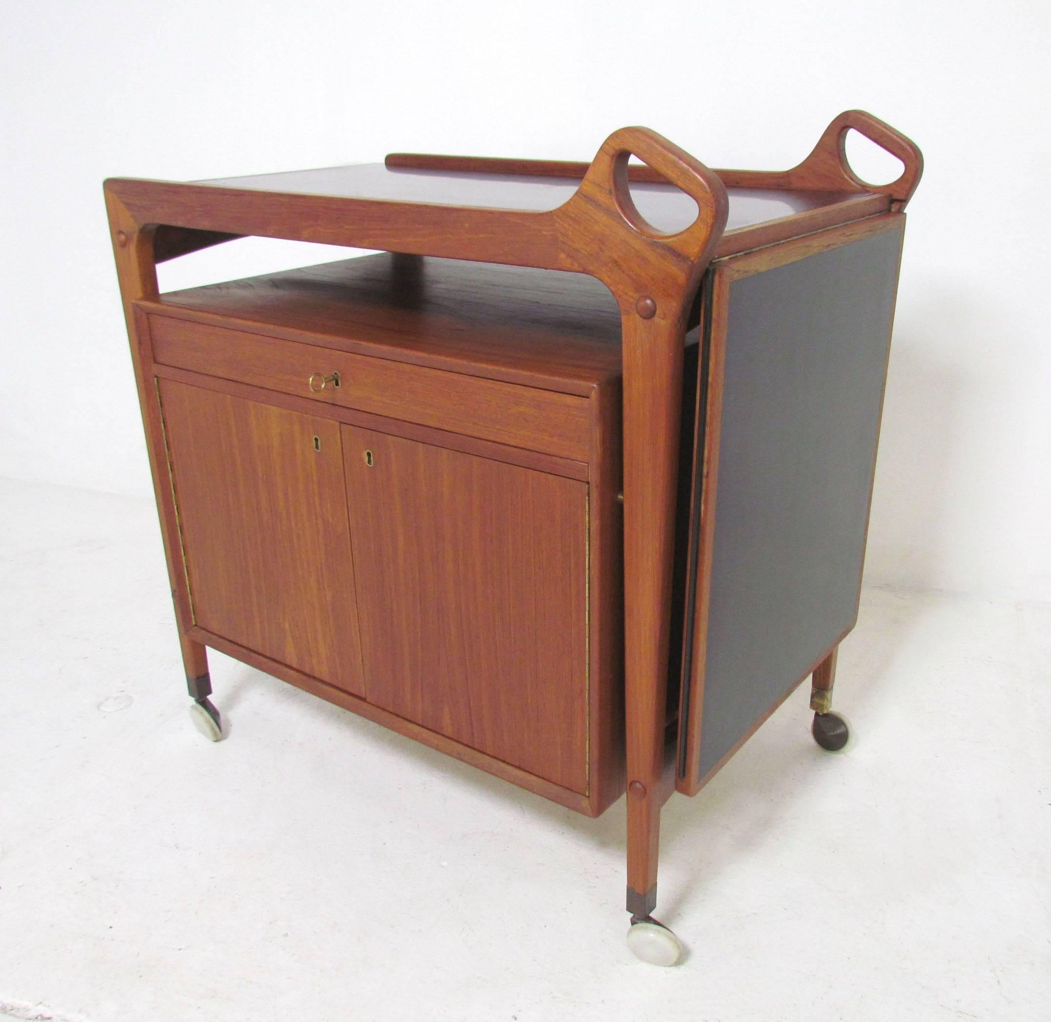 Danish teak drinks serving cart with locking storage cabinet with ample space for liquor bottles and a flatware drawer for serving utensils. Also features a spill resistant laminate top that expands if needed when drinks are being mixed and served.