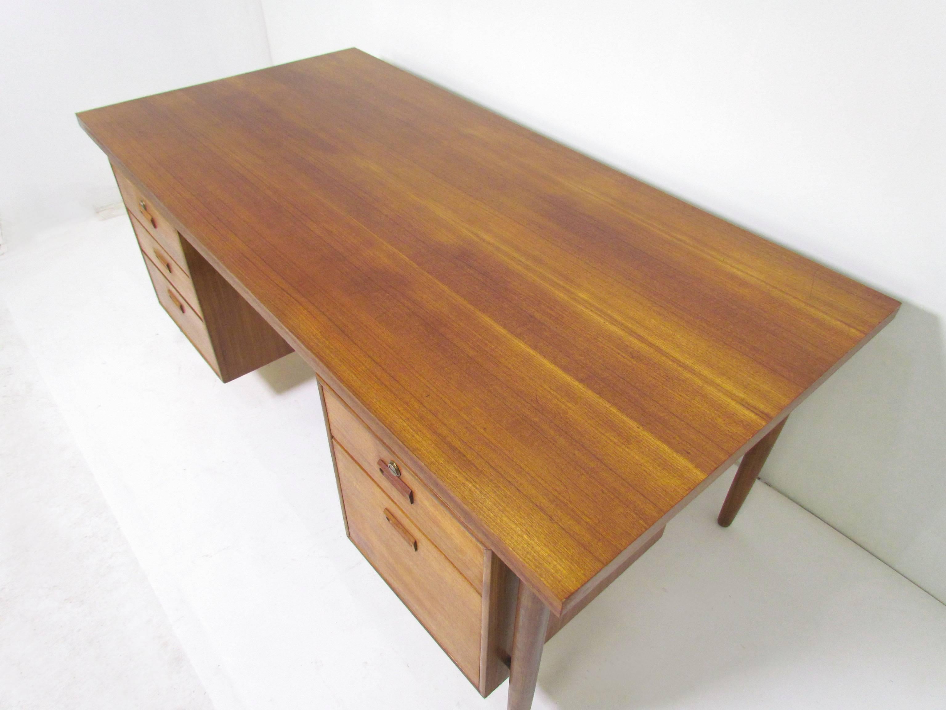 Large Mid-Century Modern executive desk in teak, circa 1960s, with flanking under cabinets, carved drawer handles, and a hanging file drawer. Finished back.