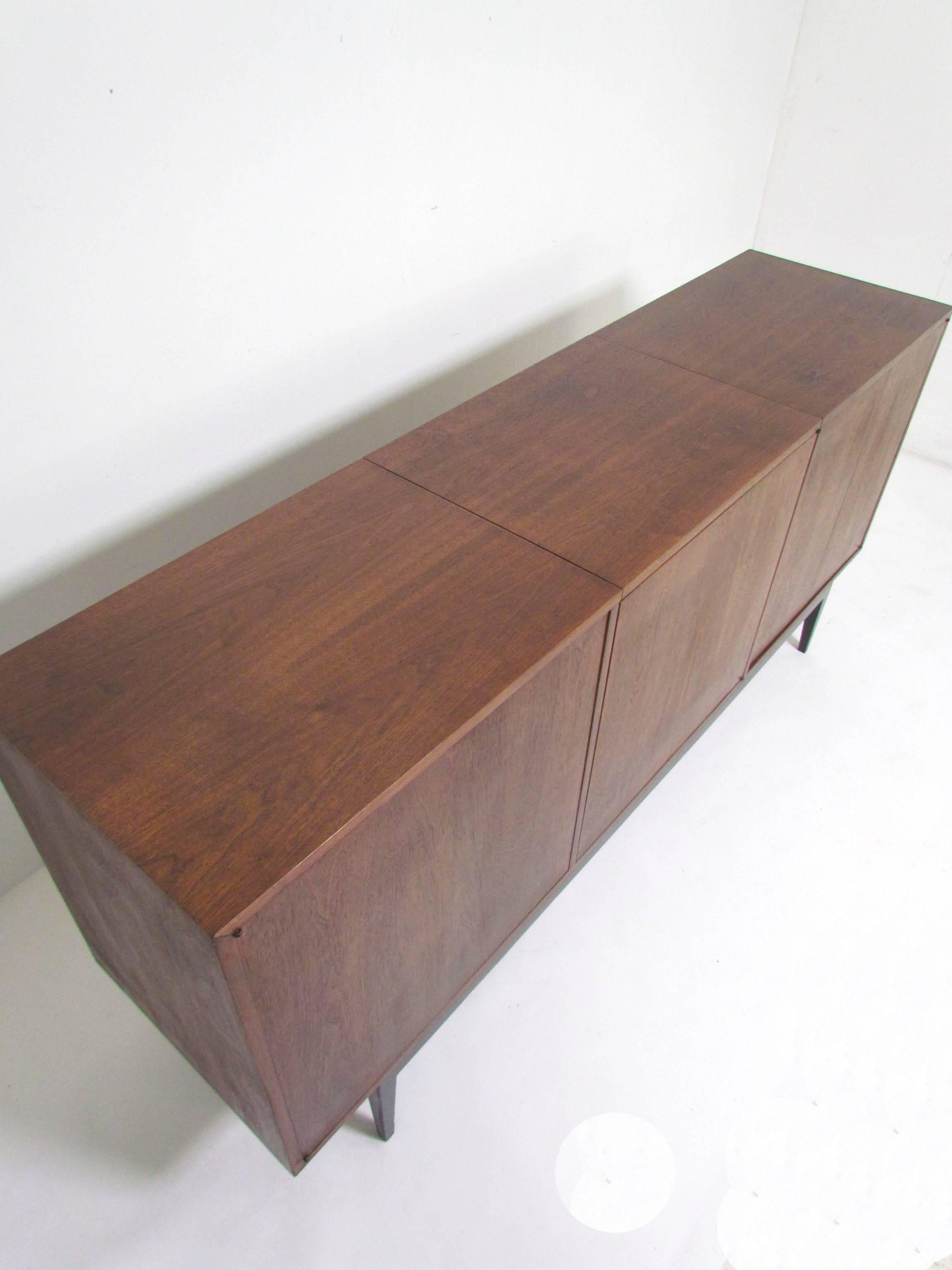 Mid-Century Modern credenza in walnut with ebonized base, three bi-fold doors reveal ample storage; top compartment reveals shallow drawer for flatware or other items. In the manner of Paul McCobb or Harvey Probber.