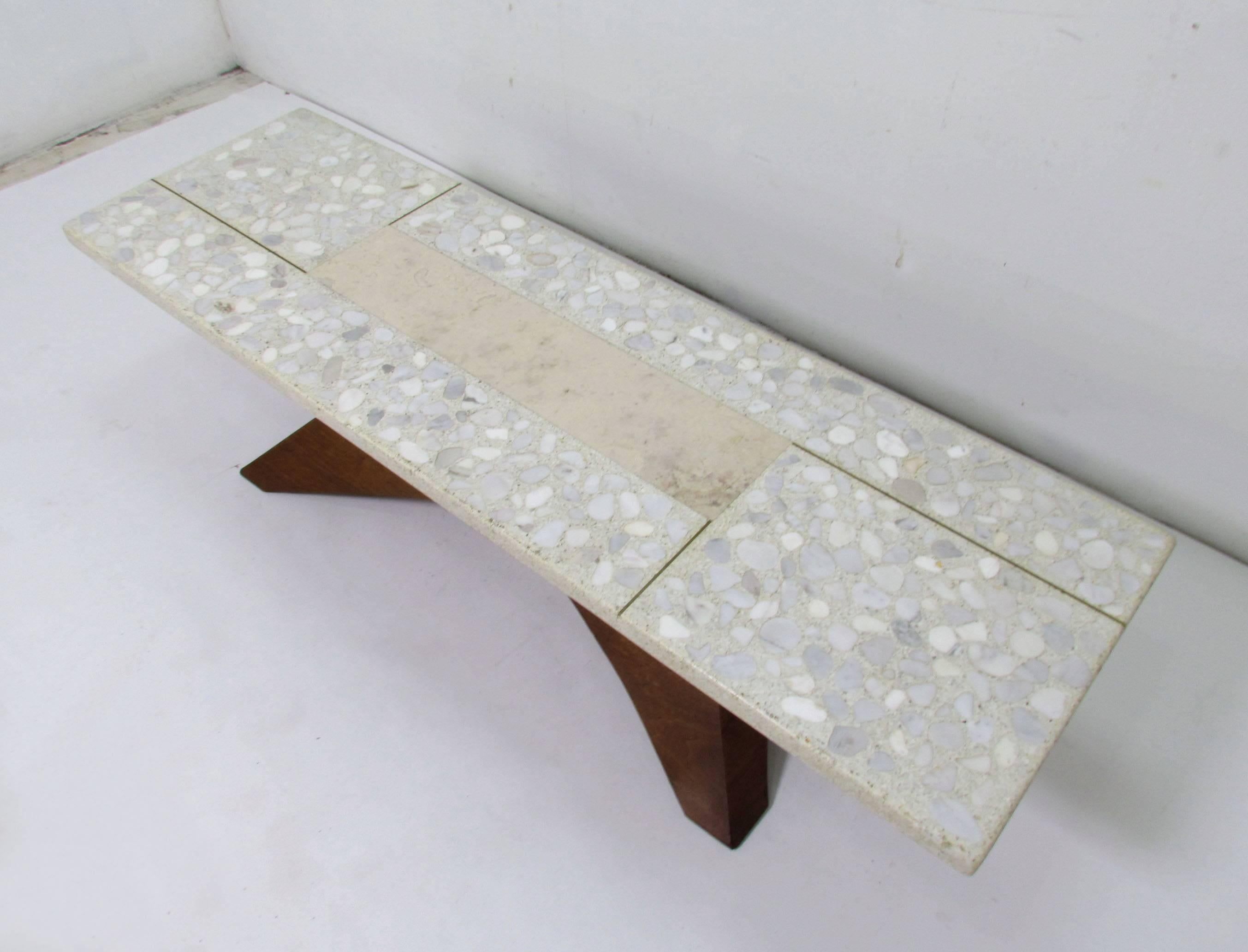Unusual form coffee table in the manner of Harvey Probber, featuring a terrazzo stone top with marble centre and brass inlay over a sculptural carved walnut base with recessed casters.