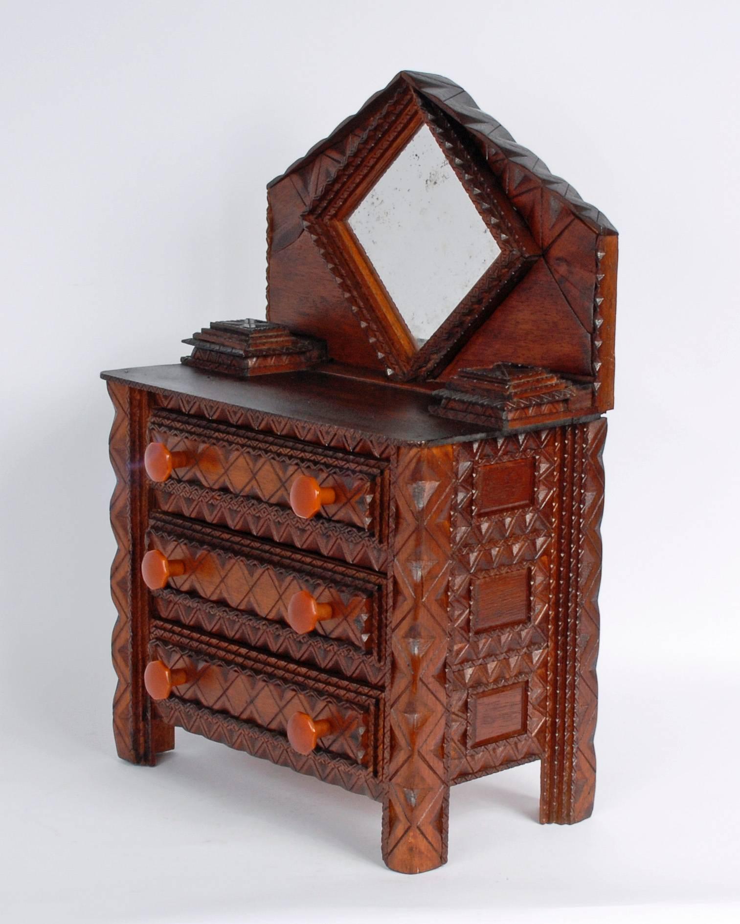 A fine Tramp Art miniature dresser with a peaked top, three large drawers with Bakelite knobs, two lift-top compartments and a diamond shaped mirror. The dresser exhibits finely carved details and the original Bakelite knobs are a nice
