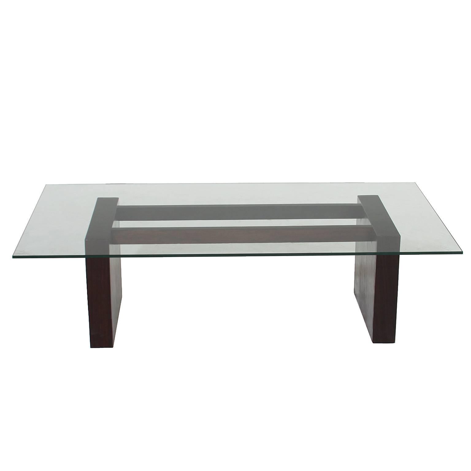 A simple coffee table with a rosewood base. The base has two stretchers along the top and two thick bases on either side of the stretcher. It creates a dynamic look to a simple coffee table. The top is clear glass. 

Many pieces are stored in our