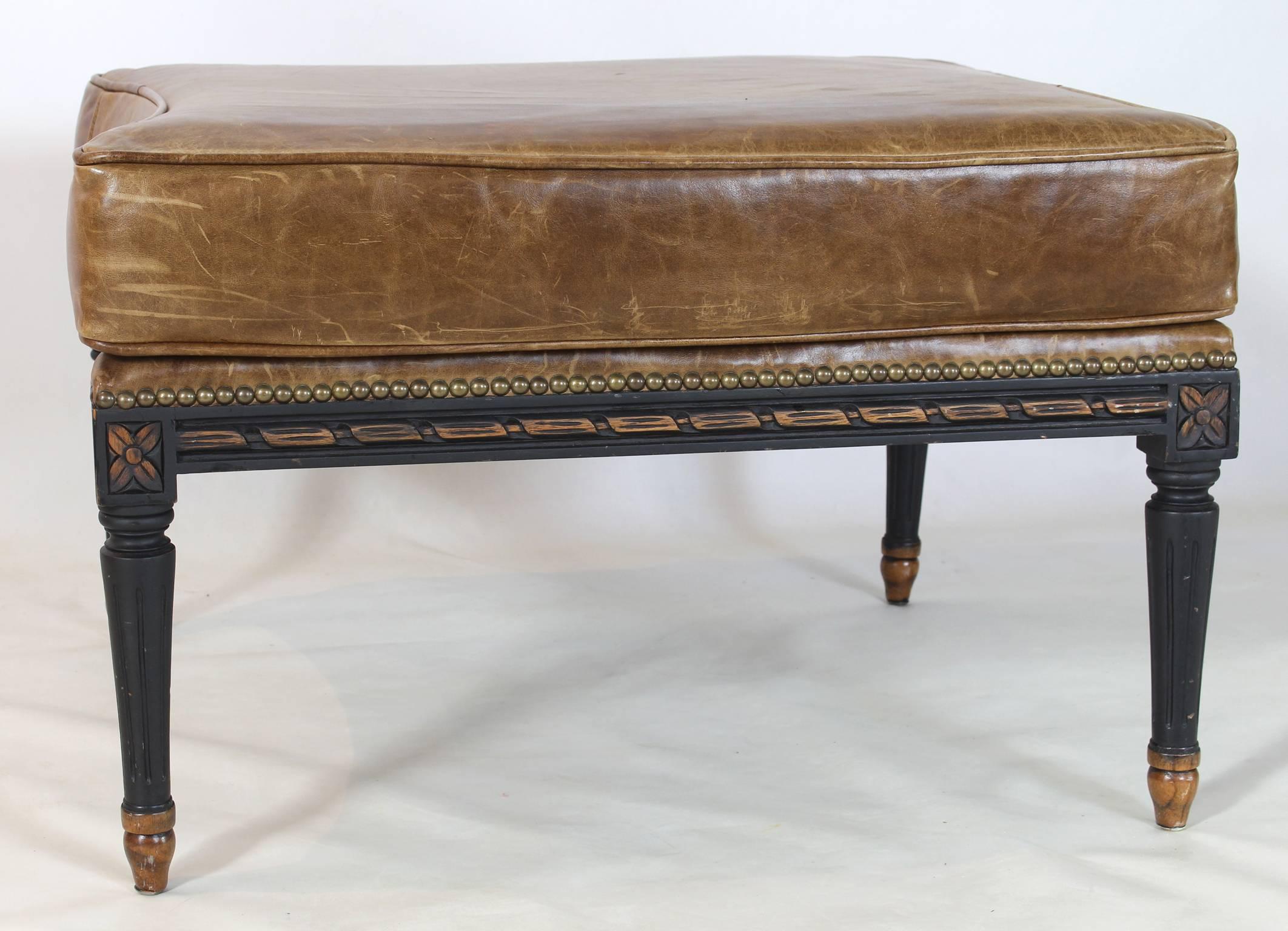 A large leather covered Regency style ottoman with carved, ebonized legs supporting a distressed leather clad pillow-form top accented with brass tacks.