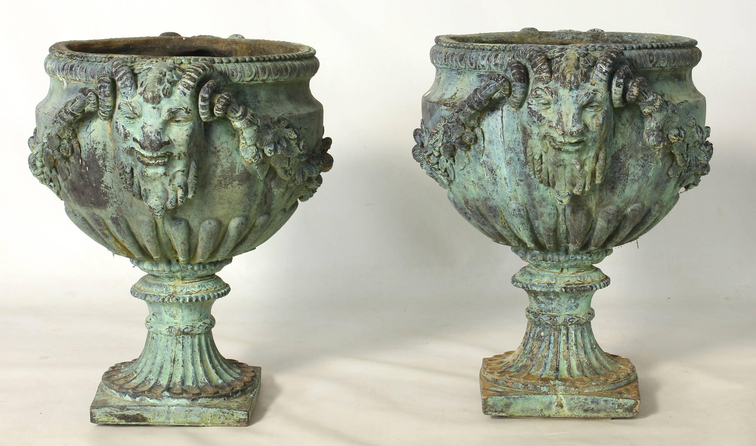 A very handsome pair of elaborately cast patinated bronze-like heavy composite urns featuring horned masks, swags and trailing vines on fluted bases.