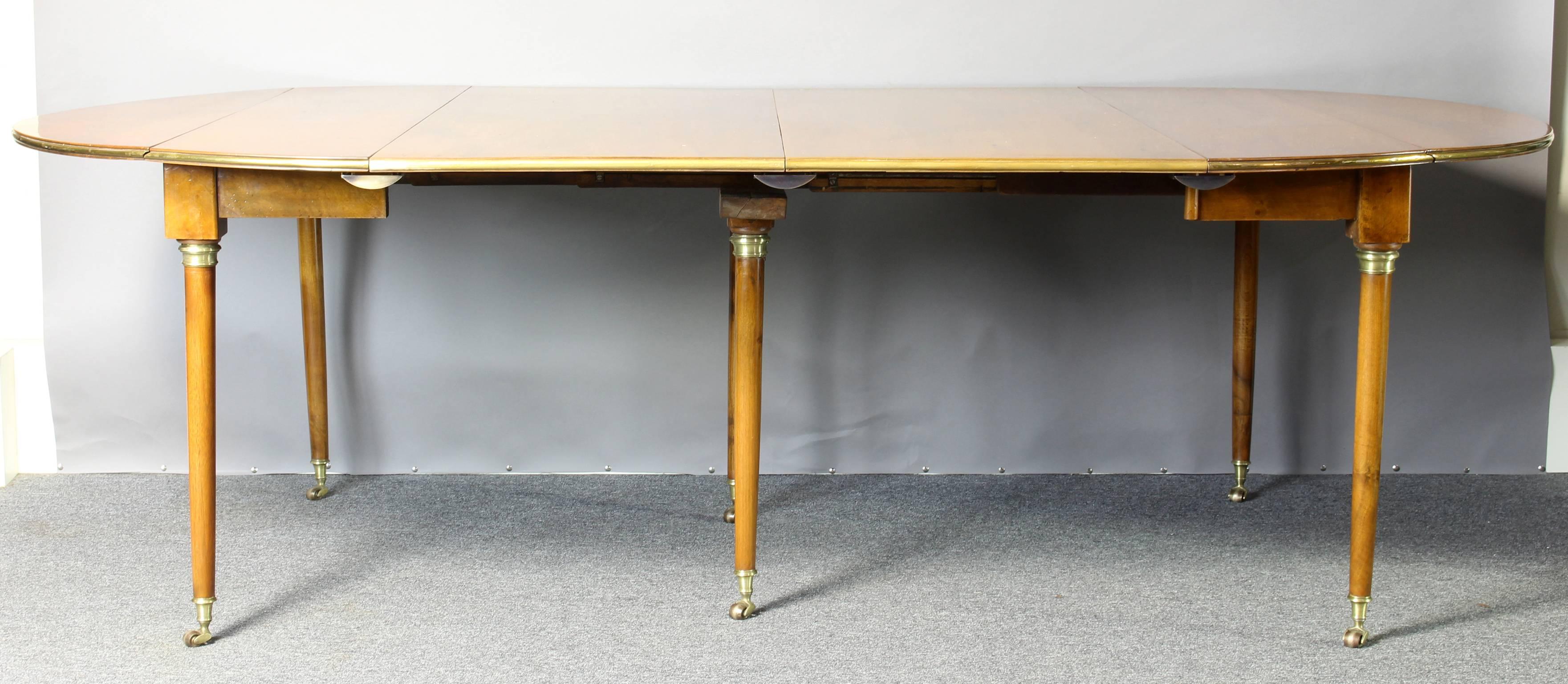 A large and elegant early 19th century French extending dining table made of a warm golden fruitwood with round tapering legs accented with brass cuffs and cup casters. This wonderfully versatile form allows for a very compact drop-leaf table