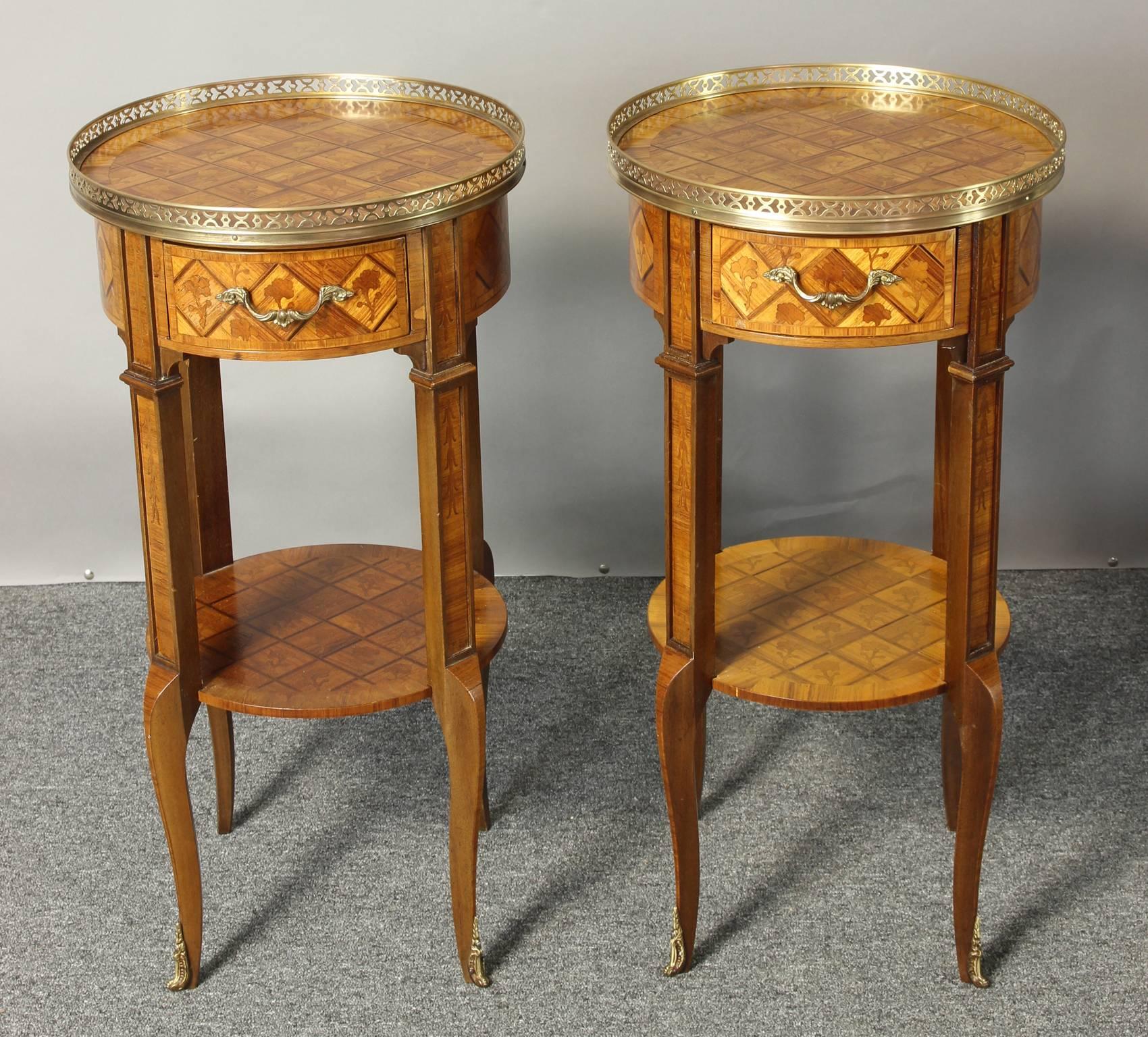An elegant pair of early 20th century French marquetry side tables in the Louis XV-XVI transition style. The top shelf and round sides have extensive marquetry with a repeating floral design and a pierced brass gallery along the top with two brass